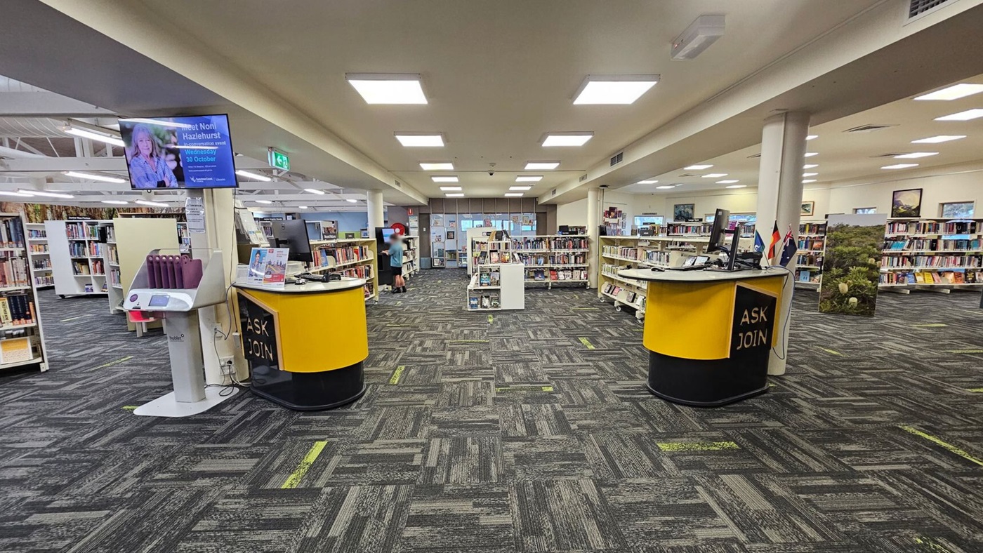 A library interior with a high, wooden ceiling supported by white beams. On the left, there are curved white bookshelves filled with books, labelled "Food and Drink." The floor is covered with dark patterned carpet tiles. On the right, there's a doorway with wooden shelving units. In the background, more bookshelves and a yellow circular desk are visible, along with a digital screen displaying content. The space is well-lit with fluorescent lights. A library interior with a high, wooden ceiling supported by white beams. On the left, there are curved white bookshelves filled with books, labelled "Food and Drink." The floor is covered with dark patterned carpet tiles. On the right, there's a doorway with wooden shelving units. In the background, more bookshelves and a yellow circular desk are visible, along with a digital screen displaying content. The space is well-lit with fluorescent lights.