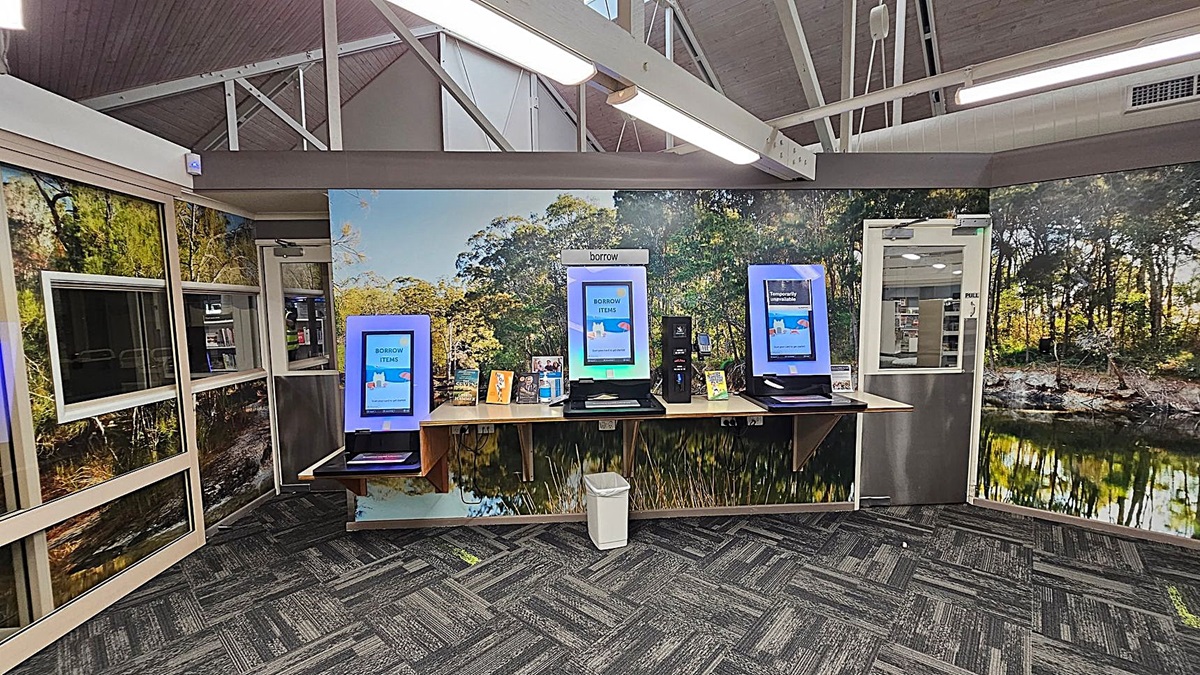 A library interior with three self-service kiosks on a wooden counter against a wall featuring a nature mural. The kiosks display screens with "BORROW ITEMS" and "Temporarily Unavailable" messages. Below the counter is a white bin. The floor is carpeted with a grey and green pattern. The ceiling has exposed beams and fluorescent lights. To the right, a glass door leads to another room with visible bookshelves. A library interior with three self-service kiosks on a wooden counter against a wall featuring a nature mural. The kiosks display screens with "BORROW ITEMS" and "Temporarily Unavailable" messages. Below the counter is a white bin. The floor is carpeted with a grey and green pattern. The ceiling has exposed beams and fluorescent lights. To the right, a glass door leads to another room with visible bookshelves.