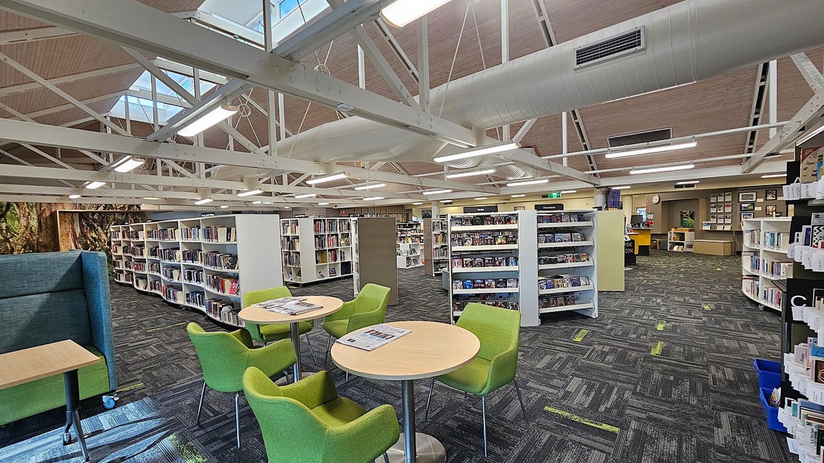 A library interior with a high ceiling featuring exposed white beams and ductwork. In the foreground, there are round tables with green chairs. The floor is carpeted in a dark pattern with green accents. Bookshelves filled with books and media are in the middle and background. On the left, there's a green cushioned seating area. The back wall has framed pictures and a clock. Skylights provide natural light from above. A library interior with a high ceiling featuring exposed white beams and ductwork. In the foreground, there are round tables with green chairs. The floor is carpeted in a dark pattern with green accents. Bookshelves filled with books and media are in the middle and background. On the left, there's a green cushioned seating area. The back wall has framed pictures and a clock. Skylights provide natural light from above.
