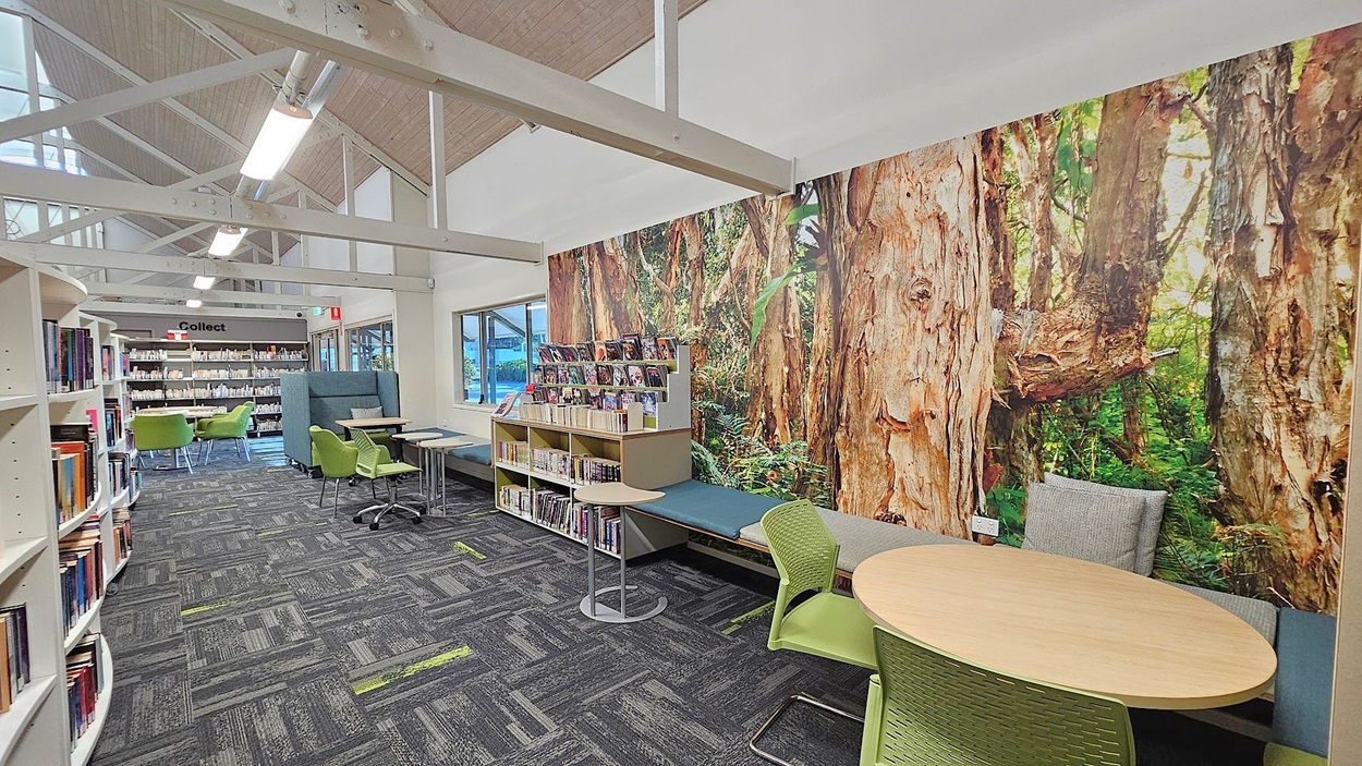 Library interior with a high, sloped ceiling featuring exposed beams. On the right, a large mural of a forest scene covers the wall, with a cushioned bench and a small round table with green chairs in front. In the middle, bookshelves display various books and magazines. On the left, more bookshelves and a seating area with green chairs and tables. The floor is carpeted with a grey and green pattern, and large windows let in natural light. Library interior with a high, sloped ceiling featuring exposed beams. On the right, a large mural of a forest scene covers the wall, with a cushioned bench and a small round table with green chairs in front. In the middle, bookshelves display various books and magazines. On the left, more bookshelves and a seating area with green chairs and tables. The floor is carpeted with a grey and green pattern, and large windows let in natural light.