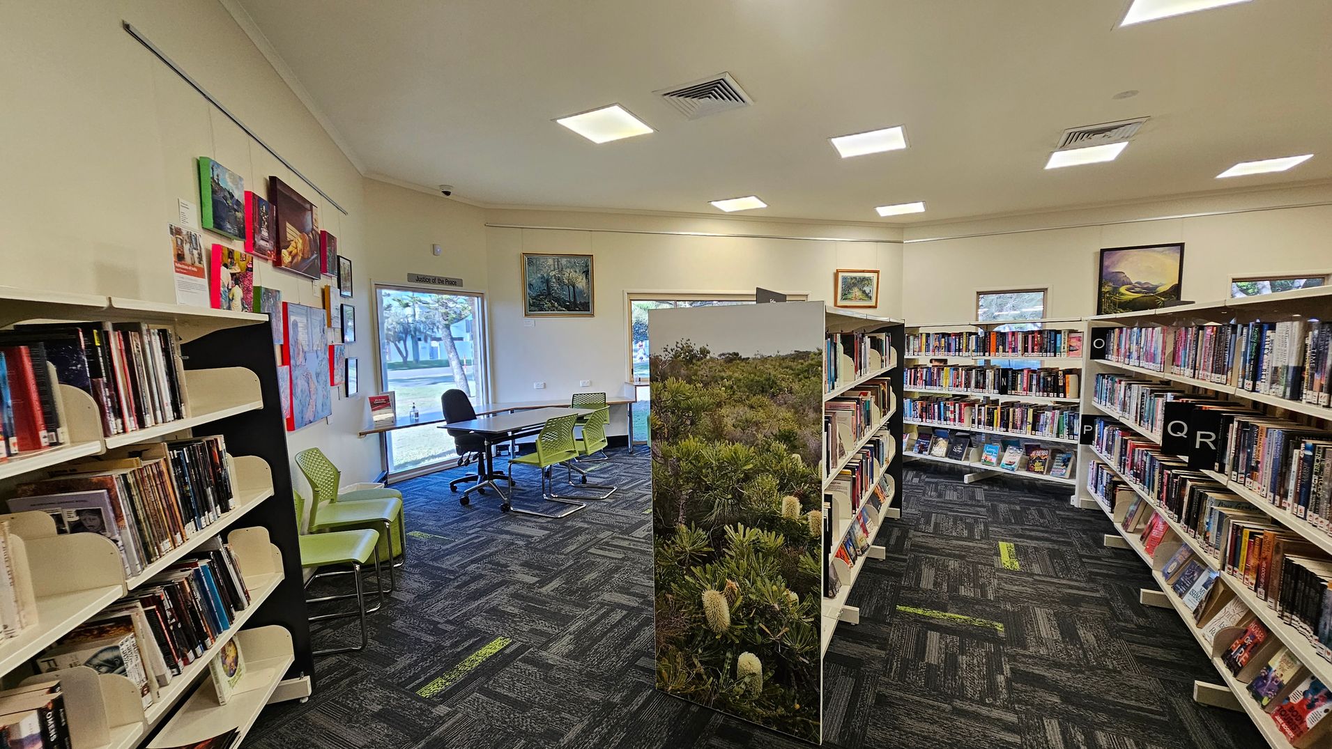 A library interior with bookshelves on the left and right, filled with books. In the middle, a partition with a nature scene separates the space. The floor is carpeted with a dark pattern featuring green accents. On the left wall, there are colourful artworks and a window with a view outside. In the background, a table with chairs is near another window. The ceiling has recessed lighting. A library interior with bookshelves on the left and right, filled with books. In the middle, a partition with a nature scene separates the space. The floor is carpeted with a dark pattern featuring green accents. On the left wall, there are colourful artworks and a window with a view outside. In the background, a table with chairs is near another window. The ceiling has recessed lighting.
