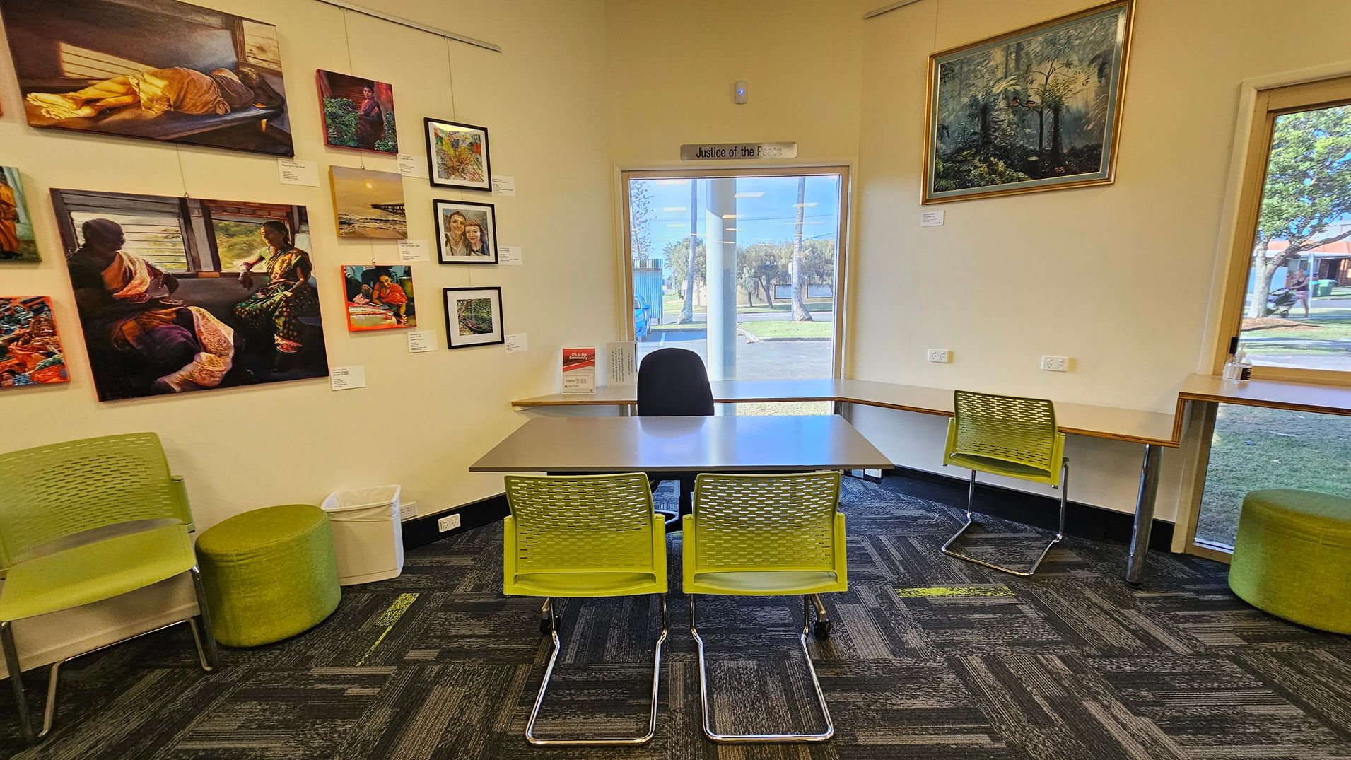 A library interior with bookshelves on both sides filled with books. In the middle, a partition displays an image of greenery. The left side has a wall with artwork and a window, next to which are tables and green chairs. The floor is carpeted with a dark pattern featuring green accents. The ceiling has recessed lighting. A library interior with bookshelves on both sides filled with books. In the middle, a partition displays an image of greenery. The left side has a wall with artwork and a window, next to which are tables and green chairs. The floor is carpeted with a dark pattern featuring green accents. The ceiling has recessed lighting.