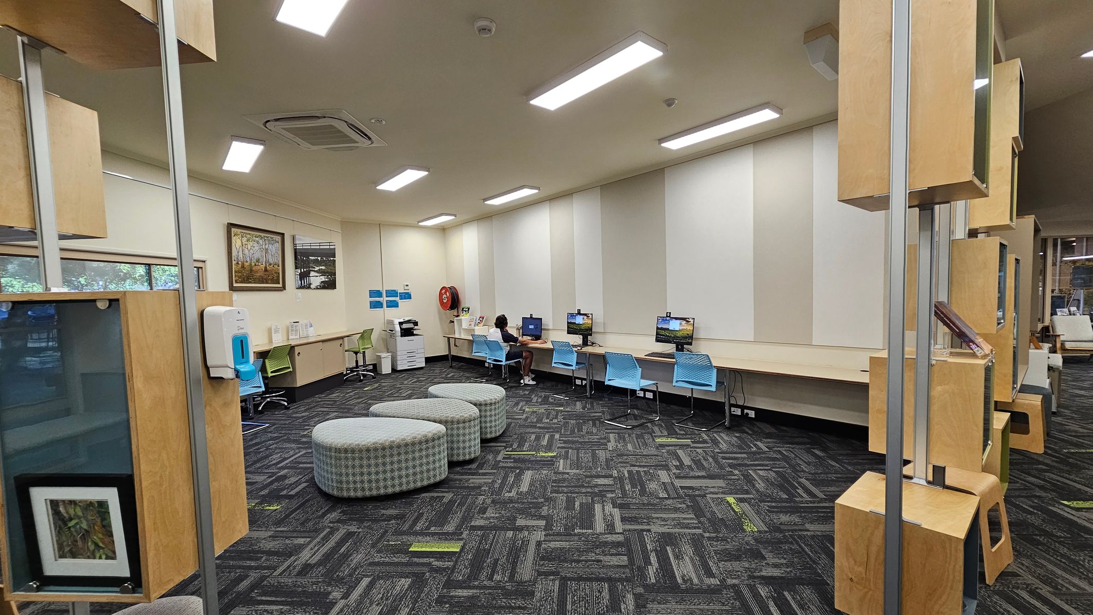 A room with a patterned carpet floor and cream walls. On the left, there are several colourful paintings and photographs displayed. Below them, a green chair and a round green ottoman are next to a white bin. In the centre, a desk with two green chairs faces a window labelled "Justice of the Peace." Behind the desk is another chair. On the right, a framed painting hangs above a long counter with a green chair. Large windows provide a view of trees and grass outside. A room with a patterned carpet floor and cream walls. On the left, there are several colourful paintings and photographs displayed. Below them, a green chair and a round green ottoman are next to a white bin. In the centre, a desk with two green chairs faces a window labelled "Justice of the Peace." Behind the desk is another chair. On the right, a framed painting hangs above a long counter with a green chair. Large windows provide a view of trees and grass outside.