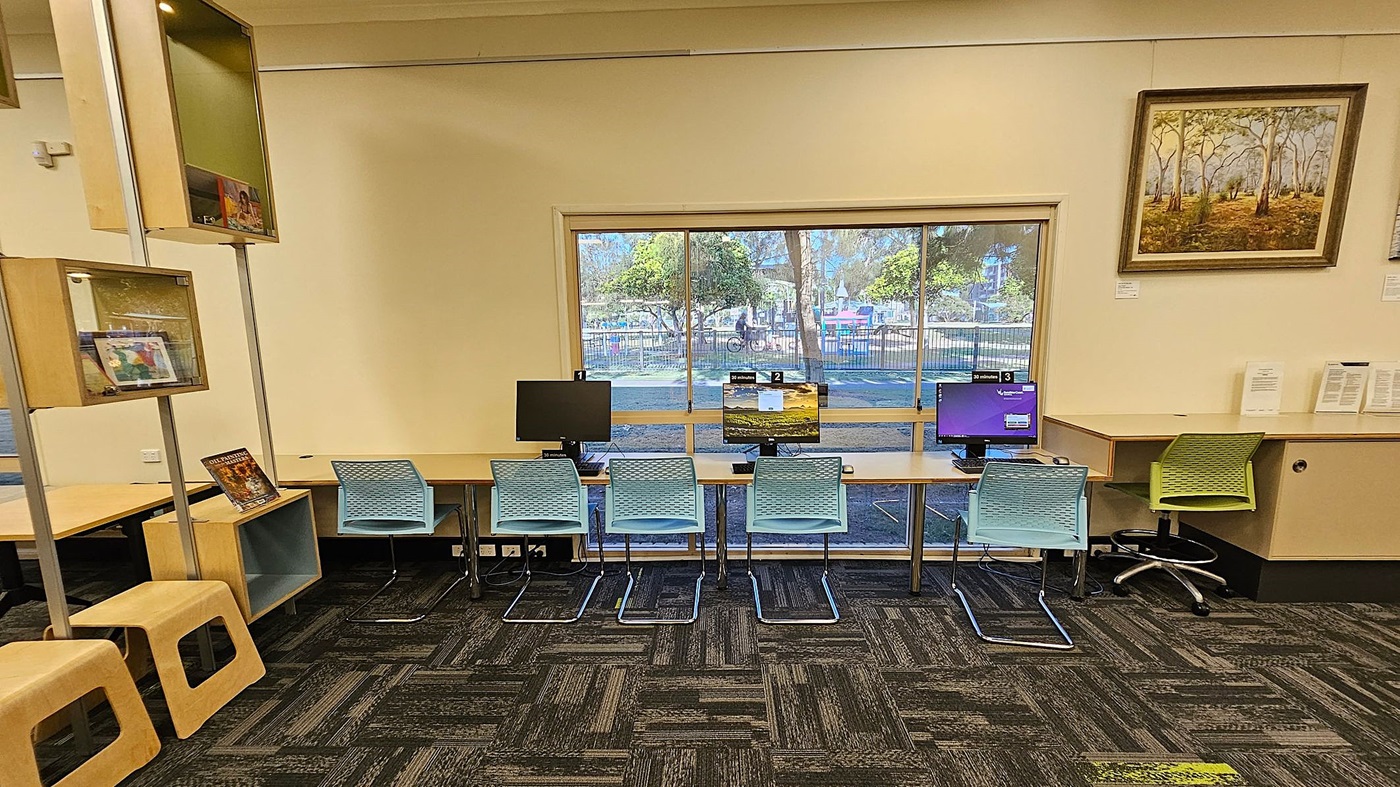 A room with a row of three computers on a long desk against a large window. Each computer has a blue chair in front of it. To the right, there's a green chair at a separate desk with papers on it. On the left, there are wooden shelves with books and a couple of wooden stools. A framed painting of trees hangs on the wall to the right. The floor is covered with a patterned carpet in shades of green and grey. A room with a row of three computers on a long desk against a large window. Each computer has a blue chair in front of it. To the right, there's a green chair at a separate desk with papers on it. On the left, there are wooden shelves with books and a couple of wooden stools. A framed painting of trees hangs on the wall to the right. The floor is covered with a patterned carpet in shades of green and grey.