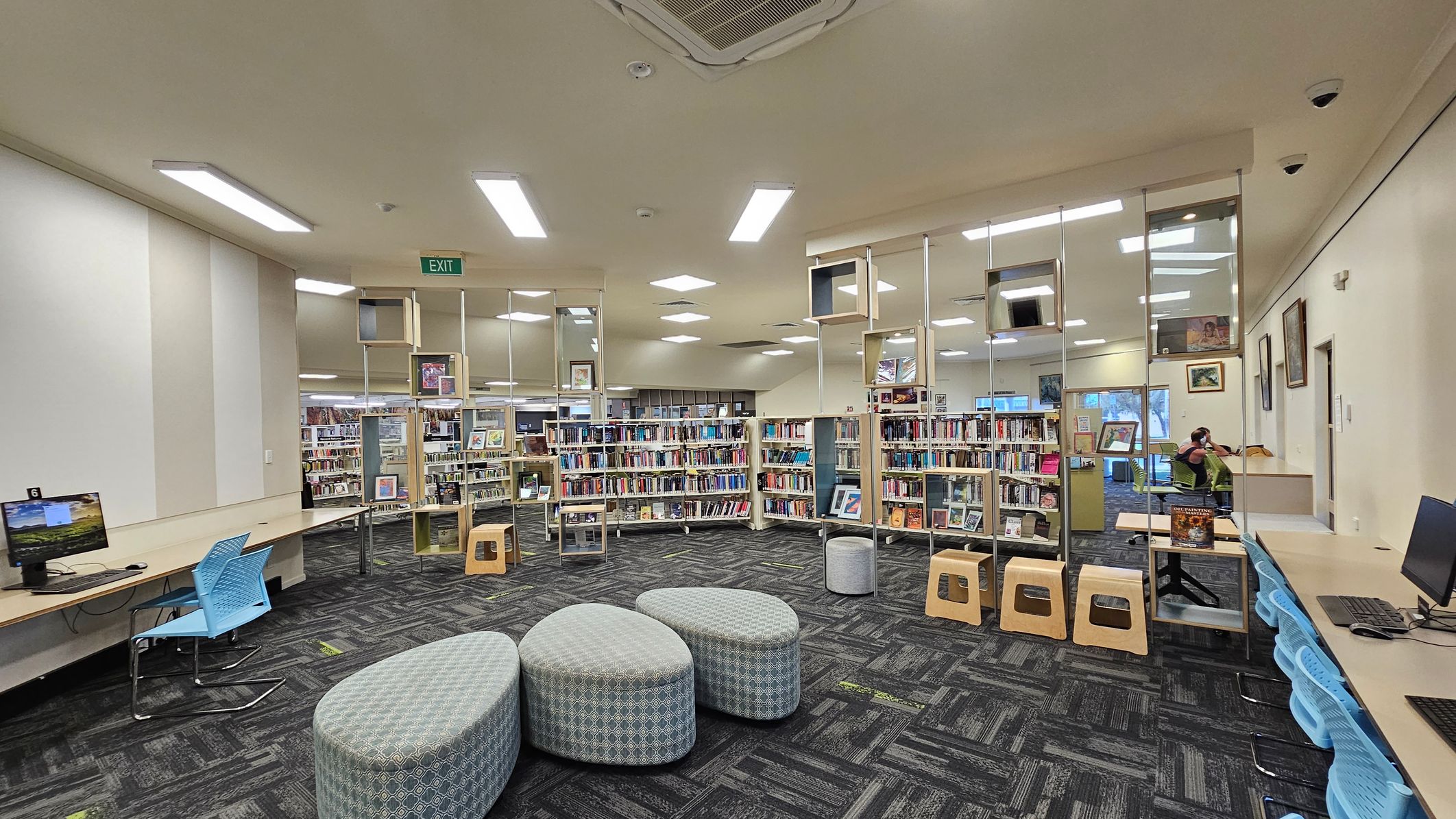 A library interior with a patterned carpet floor and a ceiling with recessed lighting. In the foreground, there are three grey, oval-shaped cushioned seats. On the left, a long desk with computers and blue chairs. In the centre, bookshelves filled with books and decorative cube shelves above. On the right, more desks with computers and a person sitting at one. The walls have framed pictures, and an exit sign is visible at the back. A library interior with a patterned carpet floor and a ceiling with recessed lighting. In the foreground, there are three grey, oval-shaped cushioned seats. On the left, a long desk with computers and blue chairs. In the centre, bookshelves filled with books and decorative cube shelves above. On the right, more desks with computers and a person sitting at one. The walls have framed pictures, and an exit sign is visible at the back.
