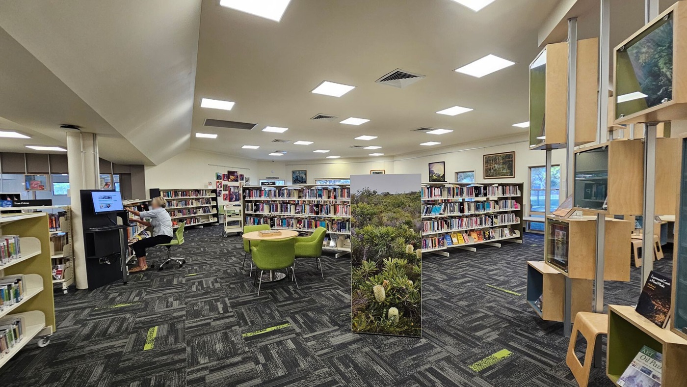 A library interior with a patterned carpet and a ceiling with square lights. On the left, a person is seated at a computer station. In the centre, there are bookshelves filled with books and a round table with green chairs. A large panel with a nature image stands nearby. On the right, there are wooden display shelves with books and framed pictures. Windows are visible in the background, allowing natural light in. A library interior with a patterned carpet and a ceiling with square lights. On the left, a person is seated at a computer station. In the centre, there are bookshelves filled with books and a round table with green chairs. A large panel with a nature image stands nearby. On the right, there are wooden display shelves with books and framed pictures. Windows are visible in the background, allowing natural light in.