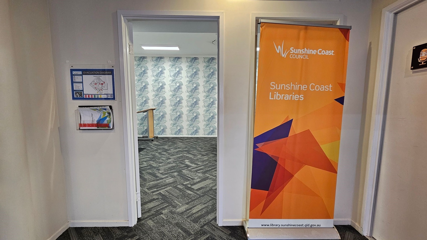 On the left, there are wooden display shelves with various items and informational signs. In the background, there are digital kiosks against a wall with a nature-themed mural. On the right, an open door leads to a room with a large flat-screen TV mounted on a white wall. The floor is covered with a patterned carpet in shades of grey and green. The ceiling is dark with recessed lighting. On the left, there are wooden display shelves with various items and informational signs. In the background, there are digital kiosks against a wall with a nature-themed mural. On the right, an open door leads to a room with a large flat-screen TV mounted on a white wall. The floor is covered with a patterned carpet in shades of grey and green. The ceiling is dark with recessed lighting.
