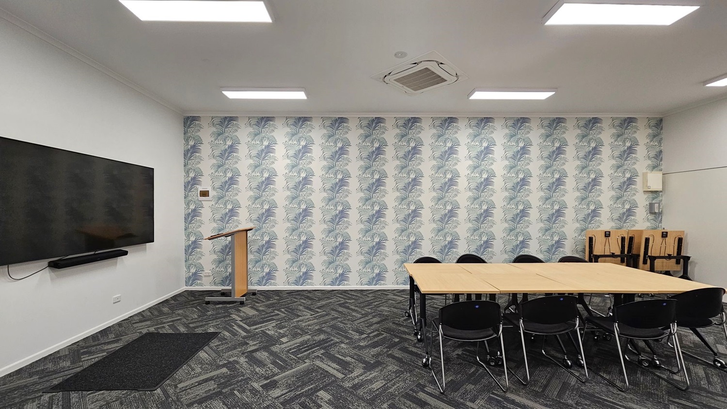 A room with a patterned wallpaper featuring leaf designs on the back wall. On the left, a large flat-screen TV is mounted on a white wall above a black mat. A wooden podium stands near the centre. On the right, a rectangular table with black chairs is arranged, and several stacked chairs are in the corner. The floor is covered with dark patterned carpet tiles, and the ceiling has recessed lighting and an air conditioning unit. A room with a patterned wallpaper featuring leaf designs on the back wall. On the left, a large flat-screen TV is mounted on a white wall above a black mat. A wooden podium stands near the centre. On the right, a rectangular table with black chairs is arranged, and several stacked chairs are in the corner. The floor is covered with dark patterned carpet tiles, and the ceiling has recessed lighting and an air conditioning unit.