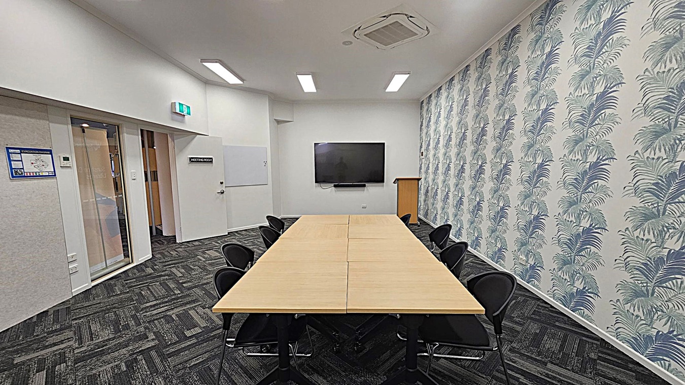 A meeting room with a rectangular wooden table in the centre, surrounded by black chairs. The right wall features a patterned wallpaper with palm leaves. At the far end, a large flat-screen TV is mounted on the wall above a wooden lectern. The left side has a door labelled "Meeting Room" and a glass panelled door. The floor is covered with dark patterned carpet tiles, and the ceiling has recessed lighting and an air conditioning unit. A meeting room with a rectangular wooden table in the centre, surrounded by black chairs. The right wall features a patterned wallpaper with palm leaves. At the far end, a large flat-screen TV is mounted on the wall above a wooden lectern. The left side has a door labelled "Meeting Room" and a glass panelled door. The floor is covered with dark patterned carpet tiles, and the ceiling has recessed lighting and an air conditioning unit.