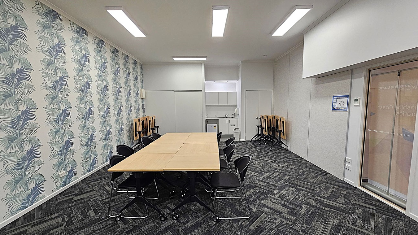 A meeting room with a long table in the centre surrounded by black chairs. On the left, the wall is decorated with a pattern of large leaves. The right side features a glass door and a plain wall with a poster. In the background, there is a small kitchenette with a sink and cabinets. The floor is covered with dark patterned carpet tiles, and the ceiling has rectangular fluorescent lights. A meeting room with a long table in the centre surrounded by black chairs. On the left, the wall is decorated with a pattern of large leaves. The right side features a glass door and a plain wall with a poster. In the background, there is a small kitchenette with a sink and cabinets. The floor is covered with dark patterned carpet tiles, and the ceiling has rectangular fluorescent lights.