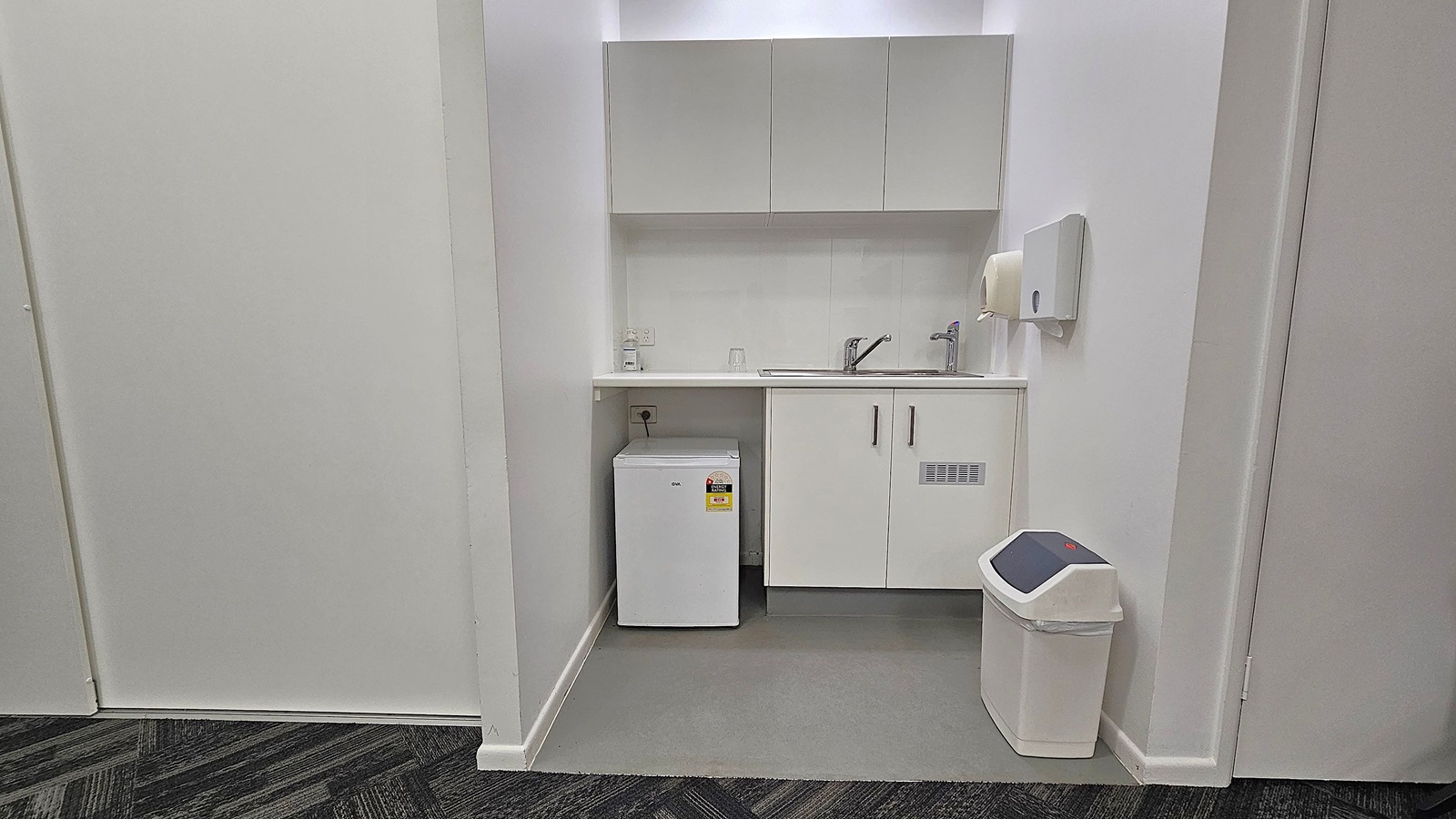 A small kitchenette with white cabinetry. On the left, there's a compact white fridge under the counter. In the centre, a sink with a tap is set into the countertop, with a soap dispenser and a glass nearby. Above, there are wall-mounted cabinets. On the right, a paper towel dispenser is mounted on the wall, and a white bin with a black lid is on the floor. The flooring is grey, and the walls are white. A small kitchenette with white cabinetry. On the left, there's a compact white fridge under the counter. In the centre, a sink with a tap is set into the countertop, with a soap dispenser and a glass nearby. Above, there are wall-mounted cabinets. On the right, a paper towel dispenser is mounted on the wall, and a white bin with a black lid is on the floor. The flooring is grey, and the walls are white.