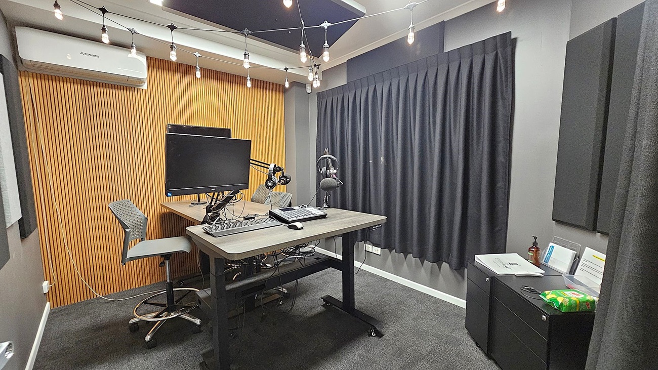 A modern recording studio with a wooden slat wall on the left and a grey curtain on the right. In the centre, there's a desk with a computer monitor, keyboard, headphones, and a microphone on an adjustable arm. Two high chairs are positioned at the desk. The ceiling has string lights hanging across. On the right, a small cabinet holds papers, a bottle, and a packet of snacks. The floor is carpeted in dark grey. A modern recording studio with a wooden slat wall on the left and a grey curtain on the right. In the centre, there's a desk with a computer monitor, keyboard, headphones, and a microphone on an adjustable arm. Two high chairs are positioned at the desk. The ceiling has string lights hanging across. On the right, a small cabinet holds papers, a bottle, and a packet of snacks. The floor is carpeted in dark grey.