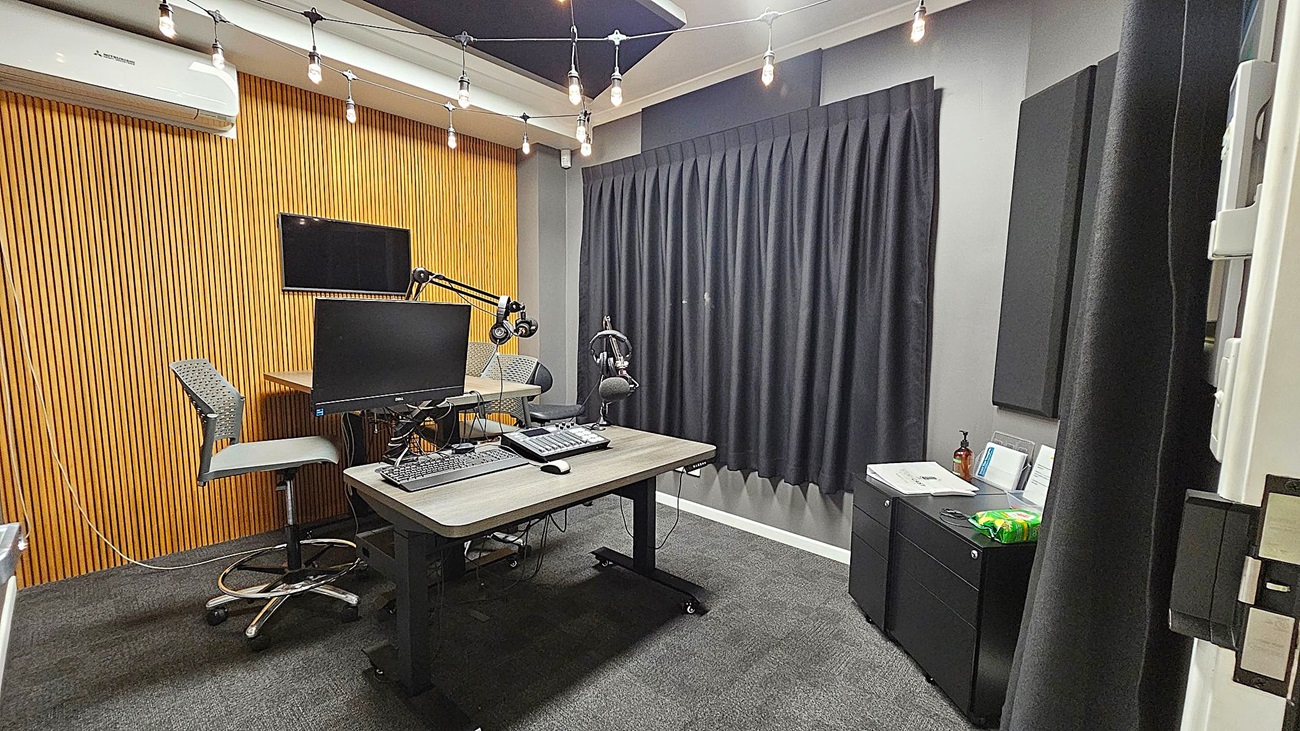 A recording studio with a wooden panelled wall on the left, featuring an air conditioning unit and a mounted monitor. Two grey chairs are positioned at a desk with a computer monitor, keyboard, and microphone. The right side has a black curtain and a small cabinet with papers and a bottle on top. The ceiling has exposed light bulbs, and the floor is carpeted in dark grey. A recording studio with a wooden panelled wall on the left, featuring an air conditioning unit and a mounted monitor. Two grey chairs are positioned at a desk with a computer monitor, keyboard, and microphone. The right side has a black curtain and a small cabinet with papers and a bottle on top. The ceiling has exposed light bulbs, and the floor is carpeted in dark grey.