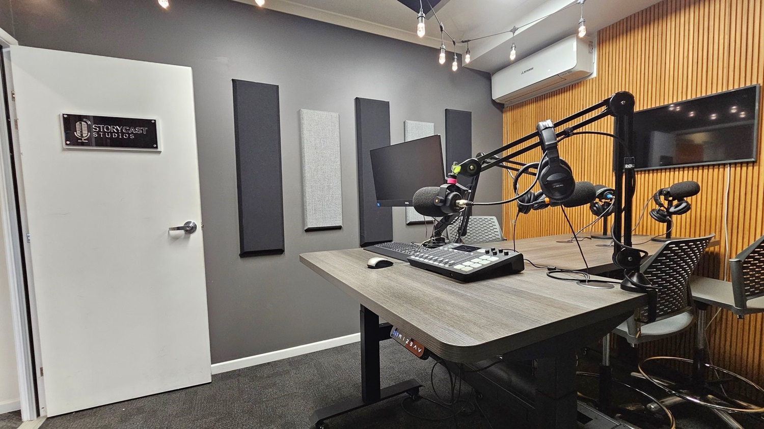 A podcast studio with a white door on the left featuring a "Storycast Studios" sign. The grey wall has acoustic panels. In the centre, a wooden table holds a computer monitor, keyboard, and audio mixer. Microphones and headphones are mounted on adjustable arms. The right wall is lined with vertical wooden slats and a flat-screen TV. The ceiling has exposed light bulbs, and the floor is carpeted in dark grey. A podcast studio with a white door on the left featuring a "Storycast Studios" sign. The grey wall has acoustic panels. In the centre, a wooden table holds a computer monitor, keyboard, and audio mixer. Microphones and headphones are mounted on adjustable arms. The right wall is lined with vertical wooden slats and a flat-screen TV. The ceiling has exposed light bulbs, and the floor is carpeted in dark grey.