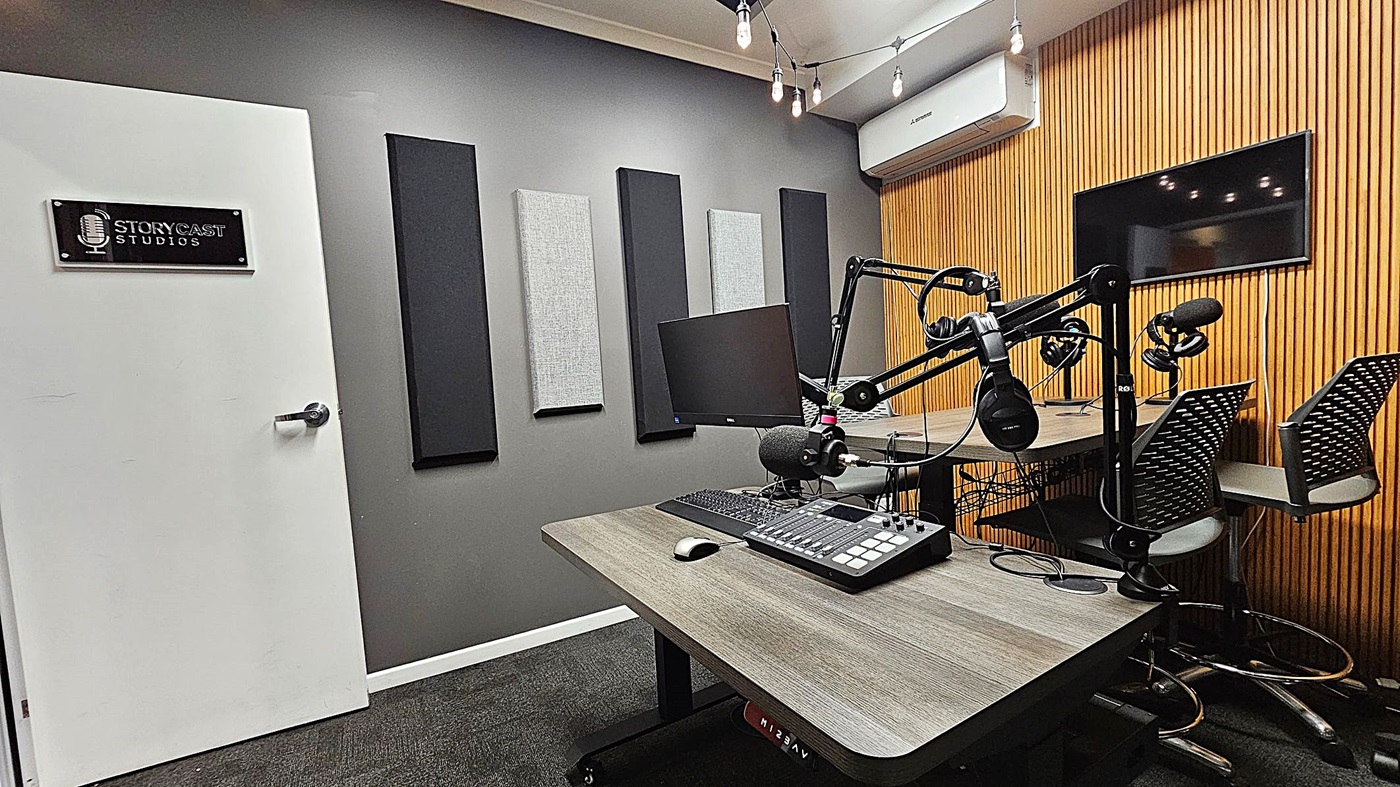 A podcast studio with a grey wall on the left featuring acoustic panels and a white door with a "Storycast Studios" sign. In the centre, there's a desk with a microphone, headphones, a computer monitor, and audio equipment. On the right, a wooden slat wall with a mounted screen and two chairs. The ceiling has hanging light bulbs, and the floor is carpeted in dark grey. An air conditioning unit is mounted above the wooden wall. A podcast studio with a grey wall on the left featuring acoustic panels and a white door with a "Storycast Studios" sign. In the centre, there's a desk with a microphone, headphones, a computer monitor, and audio equipment. On the right, a wooden slat wall with a mounted screen and two chairs. The ceiling has hanging light bulbs, and the floor is carpeted in dark grey. An air conditioning unit is mounted above the wooden wall.