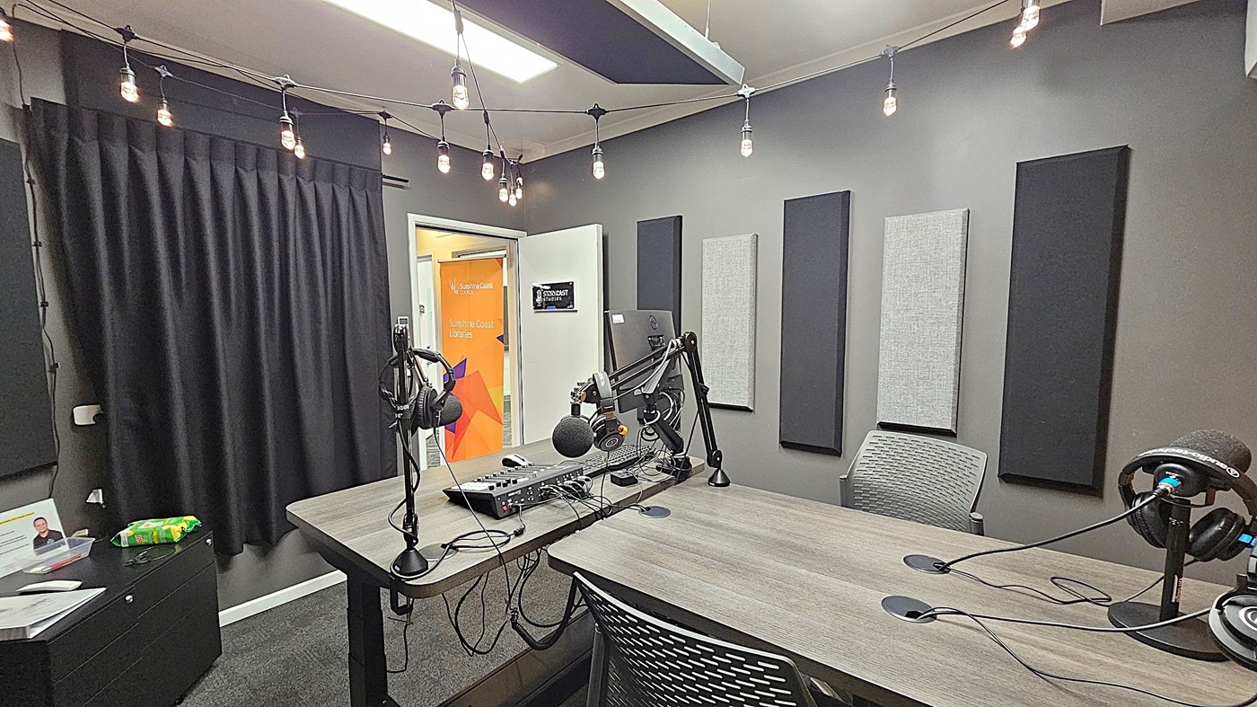 A podcast studio with a wooden table in the centre, equipped with microphones and headphones on adjustable arms. The walls are grey with acoustic panels. A string of exposed bulbs hangs from the ceiling. On the left, a black curtain covers part of the wall, and a small cabinet holds some items. In the background, a door is open, revealing a colourful sign with text. The floor is carpeted in dark grey. A podcast studio with a wooden table in the centre, equipped with microphones and headphones on adjustable arms. The walls are grey with acoustic panels. A string of exposed bulbs hangs from the ceiling. On the left, a black curtain covers part of the wall, and a small cabinet holds some items. In the background, a door is open, revealing a colourful sign with text. The floor is carpeted in dark grey.