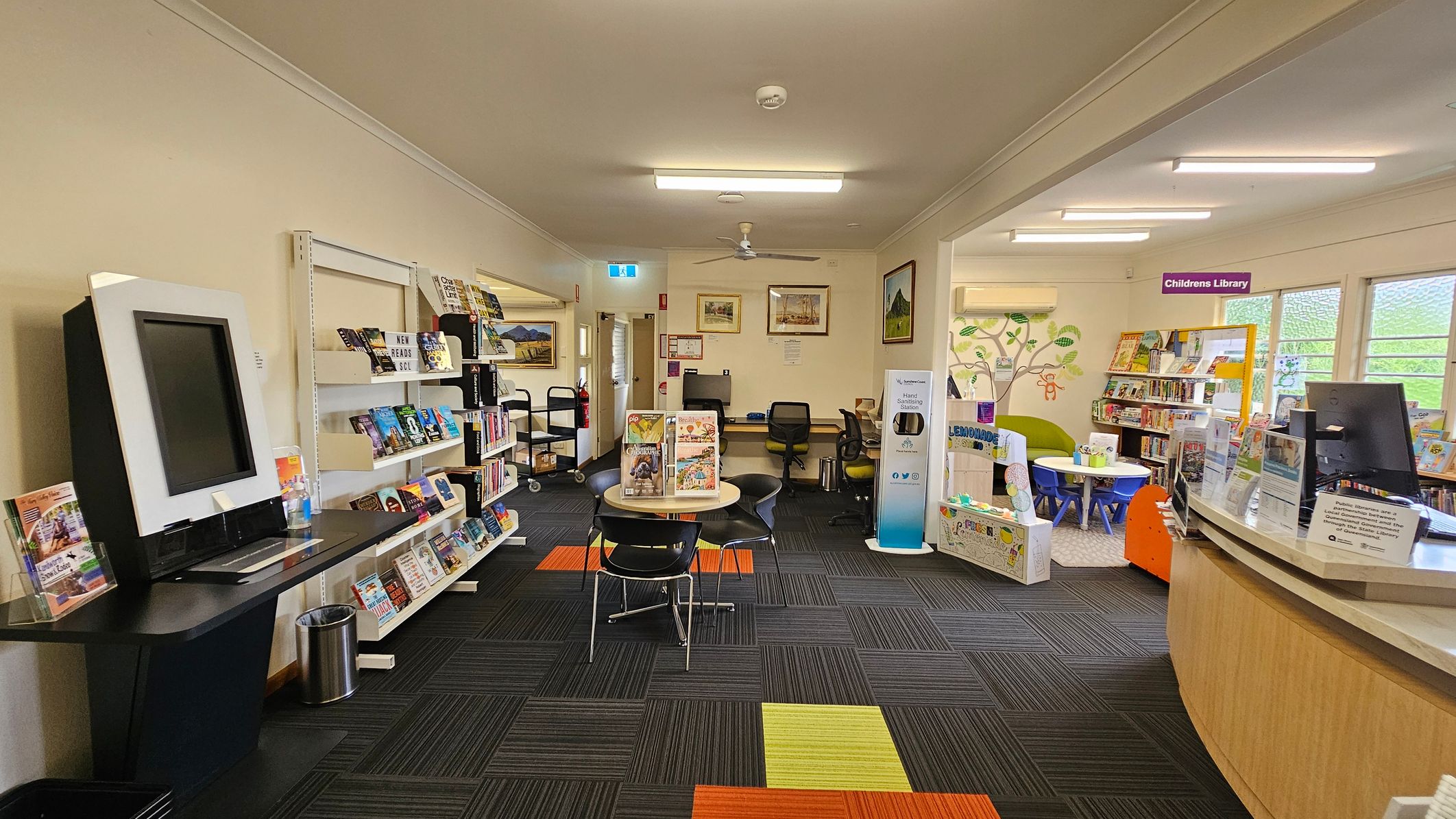 A library interior with a view through an open door. On the left, there are bookshelves filled with books and a poster on the wall. In the centre, a small round table with chairs is placed on a carpet with colourful square patterns. At the back, there are two computer stations with chairs. On the right, a glass partition displays more posters and a glimpse into another room with additional bookshelves and a counter. The ceiling has fluorescent lights. A library interior with a view through an open door. On the left, there are bookshelves filled with books and a poster on the wall. In the centre, a small round table with chairs is placed on a carpet with colourful square patterns. At the back, there are two computer stations with chairs. On the right, a glass partition displays more posters and a glimpse into another room with additional bookshelves and a counter. The ceiling has fluorescent lights.