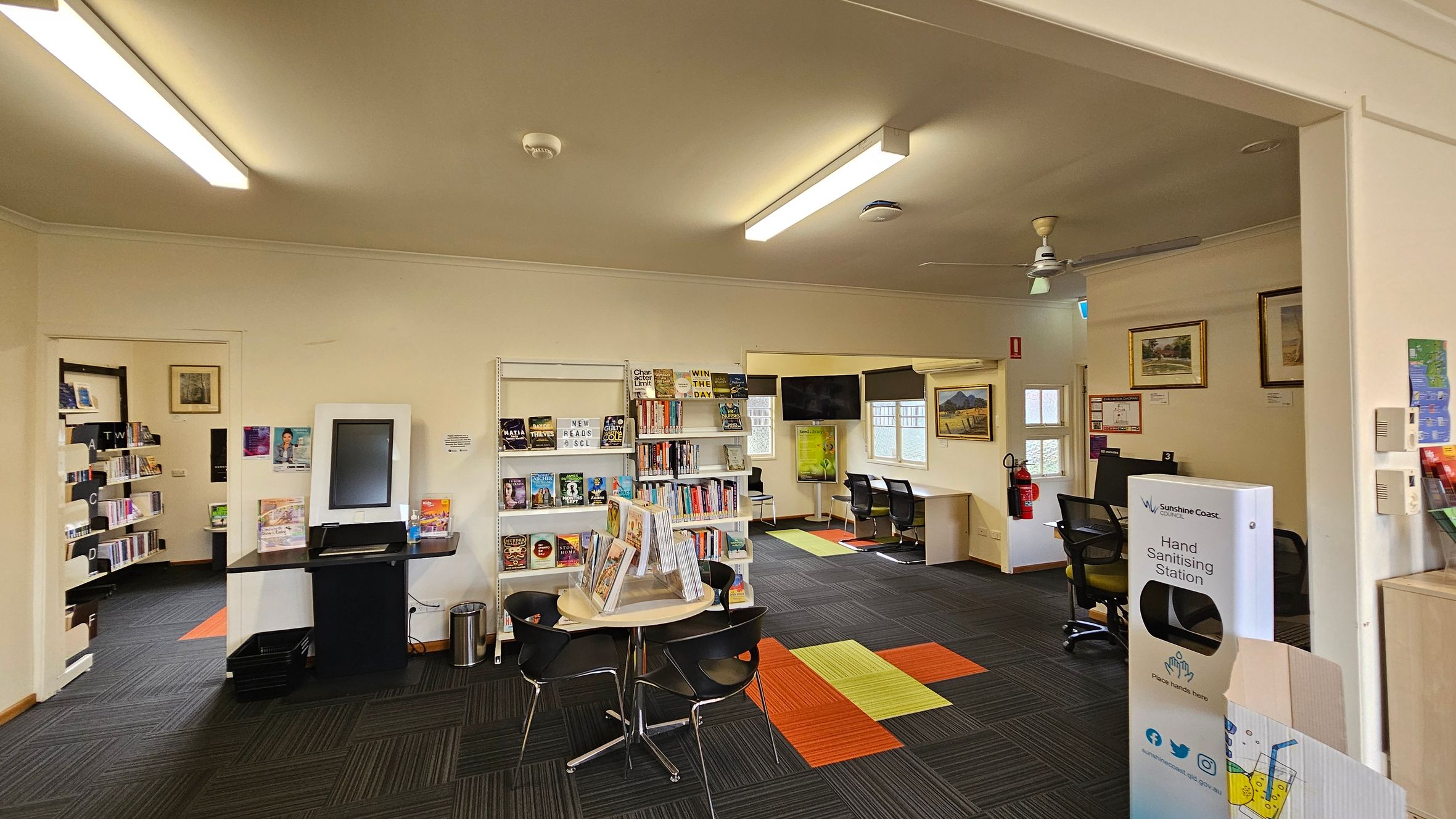 A library interior with a modern design. On the left, there are bookshelves filled with books and a digital kiosk. In the centre, a round table with chairs and a display of books. The right side features a hand sanitising station and framed artwork on the walls. The floor has a patterned carpet with green, orange, and black squares. The ceiling has fluorescent lights and a ceiling fan. In the background, there are windows and a small seating area with chairs and a table. A library interior with a modern design. On the left, there are bookshelves filled with books and a digital kiosk. In the centre, a round table with chairs and a display of books. The right side features a hand sanitising station and framed artwork on the walls. The floor has a patterned carpet with green, orange, and black squares. The ceiling has fluorescent lights and a ceiling fan. In the background, there are windows and a small seating area with chairs and a table.
