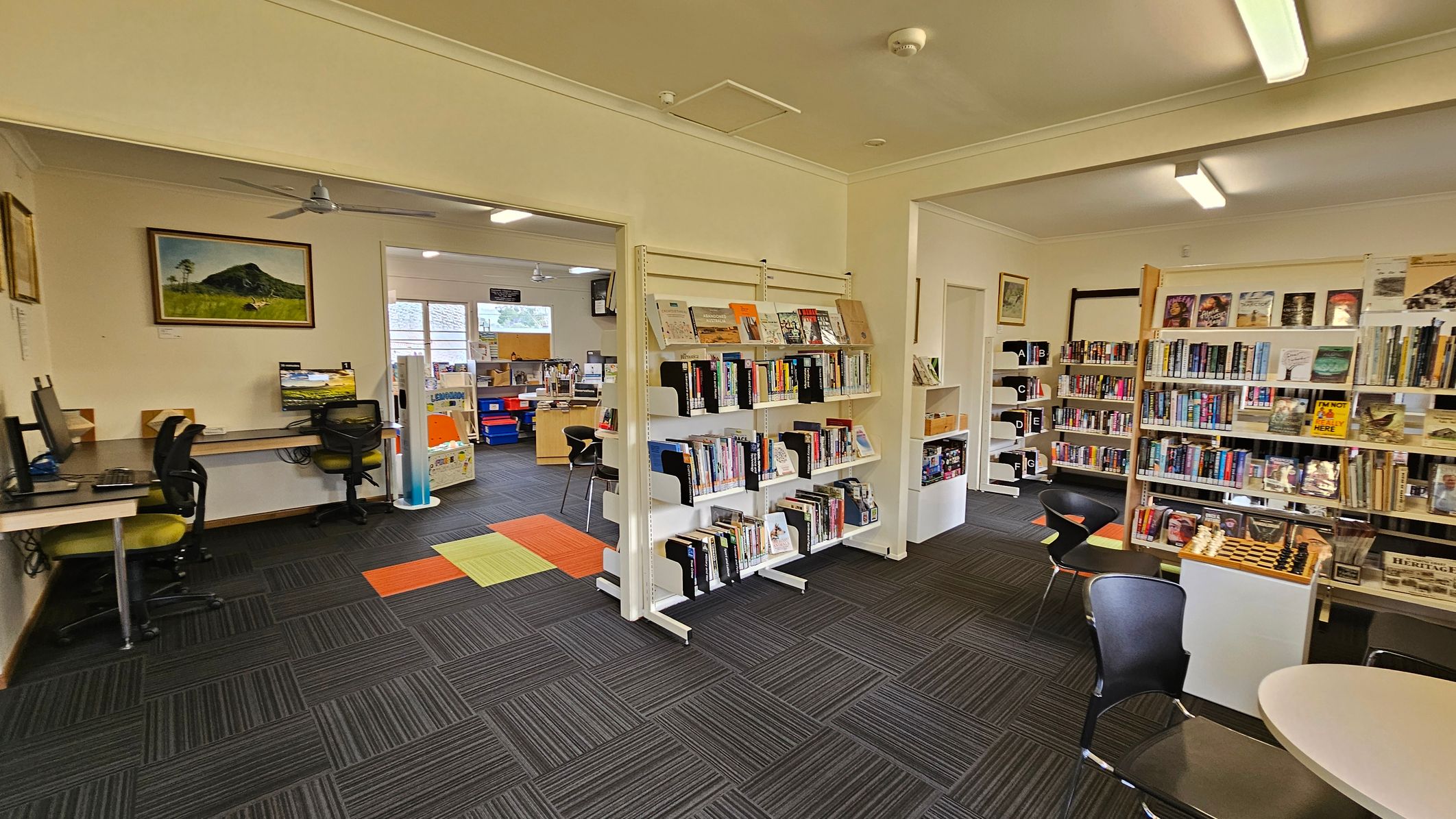 A library interior with bookshelves filled with books on the right. In the centre, a white shelving unit displays more books. On the left, there are computer workstations with chairs. A painting of a mountain hangs on the wall above the computers. The floor is covered with dark grey carpet tiles, accented with orange and green tiles. The ceiling has fluorescent lights and a ceiling fan. In the background, there is a window and more bookshelves. A library interior with bookshelves filled with books on the right. In the centre, a white shelving unit displays more books. On the left, there are computer workstations with chairs. A painting of a mountain hangs on the wall above the computers. The floor is covered with dark grey carpet tiles, accented with orange and green tiles. The ceiling has fluorescent lights and a ceiling fan. In the background, there is a window and more bookshelves.