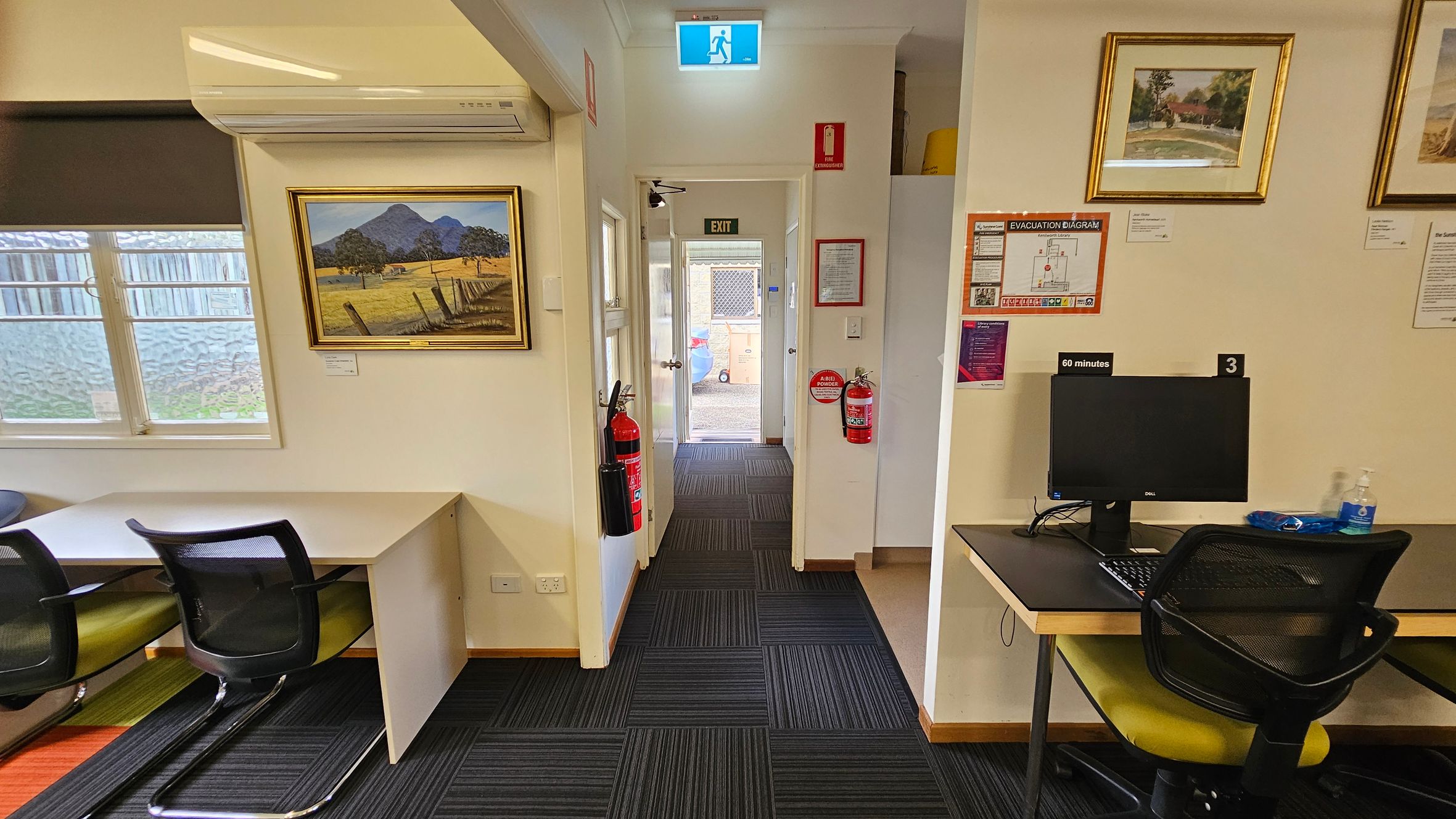 A room with a black and grey striped carpet. On the left, there's a window with a blind, a desk, and a chair. Above the desk is an air conditioning unit and a framed landscape painting. In the centre, a hallway leads to an exit door with a lit exit sign above. Fire extinguishers are mounted on the walls. On the right, there's a computer on a desk with a chair, and framed pictures on the wall. An evacuation diagram and other notices are displayed nearby. A room with a black and grey striped carpet. On the left, there's a window with a blind, a desk, and a chair. Above the desk is an air conditioning unit and a framed landscape painting. In the centre, a hallway leads to an exit door with a lit exit sign above. Fire extinguishers are mounted on the walls. On the right, there's a computer on a desk with a chair, and framed pictures on the wall. An evacuation diagram and other notices are displayed nearby.