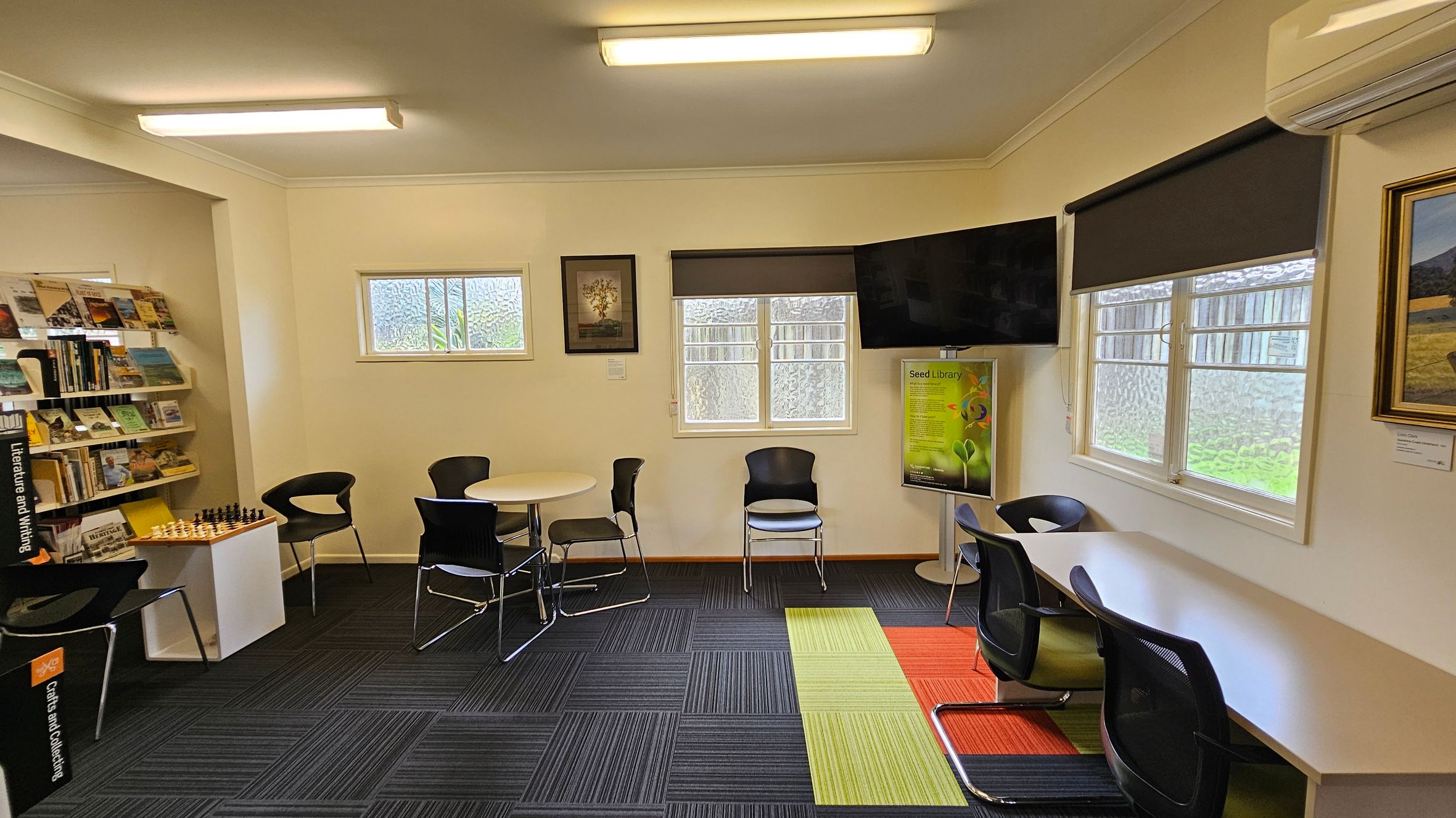 A library room with a modern design. On the left, there are bookshelves filled with books and a chess set on a small table. In the centre, a round table with black chairs is placed near two small windows. A framed picture hangs between the windows. On the right, a long desk with chairs is positioned under another window. A large TV is mounted above a poster stand. The floor has a mix of dark, green, and orange carpet tiles, and the ceiling has fluorescent lights. A library room with a modern design. On the left, there are bookshelves filled with books and a chess set on a small table. In the centre, a round table with black chairs is placed near two small windows. A framed picture hangs between the windows. On the right, a long desk with chairs is positioned under another window. A large TV is mounted above a poster stand. The floor has a mix of dark, green, and orange carpet tiles, and the ceiling has fluorescent lights.