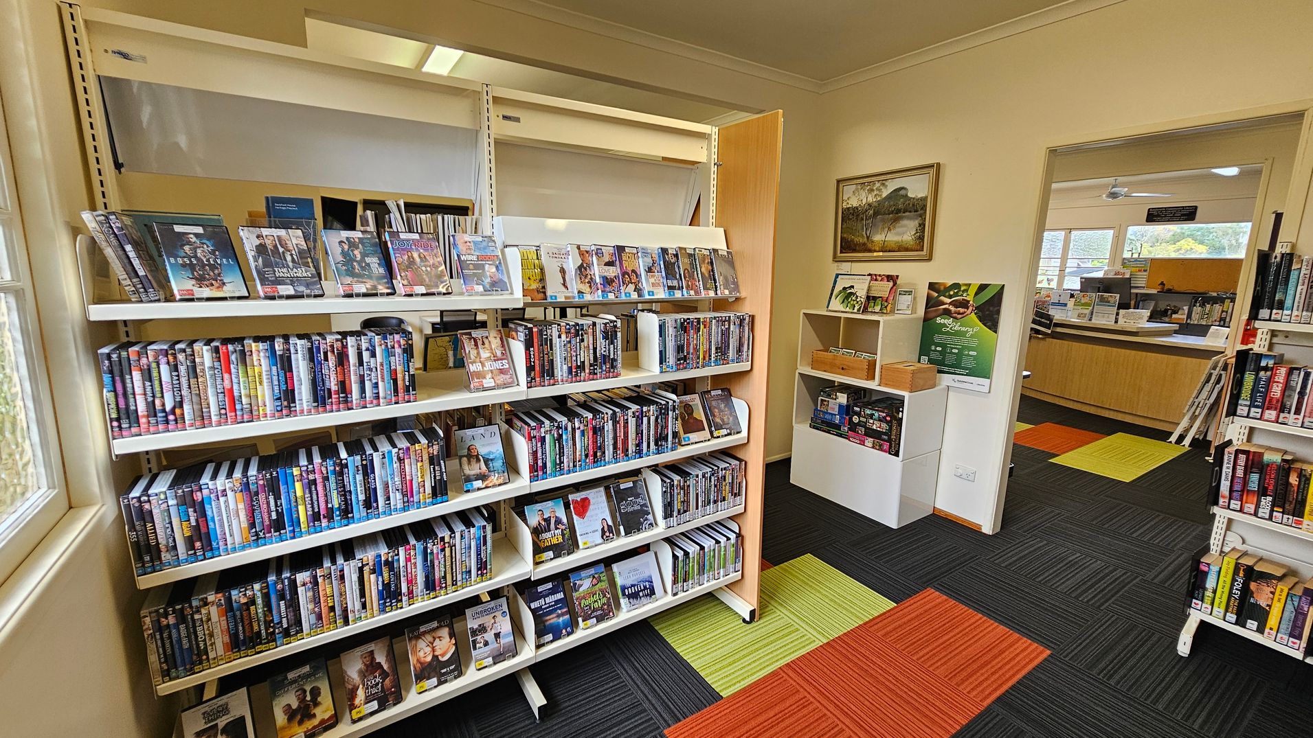 A library interior with shelves filled with DVDs on the left. The DVDs are arranged in rows, with some displayed facing forward. To the right, there's a small white shelf with brochures and a poster on the wall above it. An open doorway leads to another room with a wooden counter and large windows. The floor is covered with carpet tiles in black, green, and orange. A framed landscape painting hangs on the wall near the doorway. A library interior with shelves filled with DVDs on the left. The DVDs are arranged in rows, with some displayed facing forward. To the right, there's a small white shelf with brochures and a poster on the wall above it. An open doorway leads to another room with a wooden counter and large windows. The floor is covered with carpet tiles in black, green, and orange. A framed landscape painting hangs on the wall near the doorway.