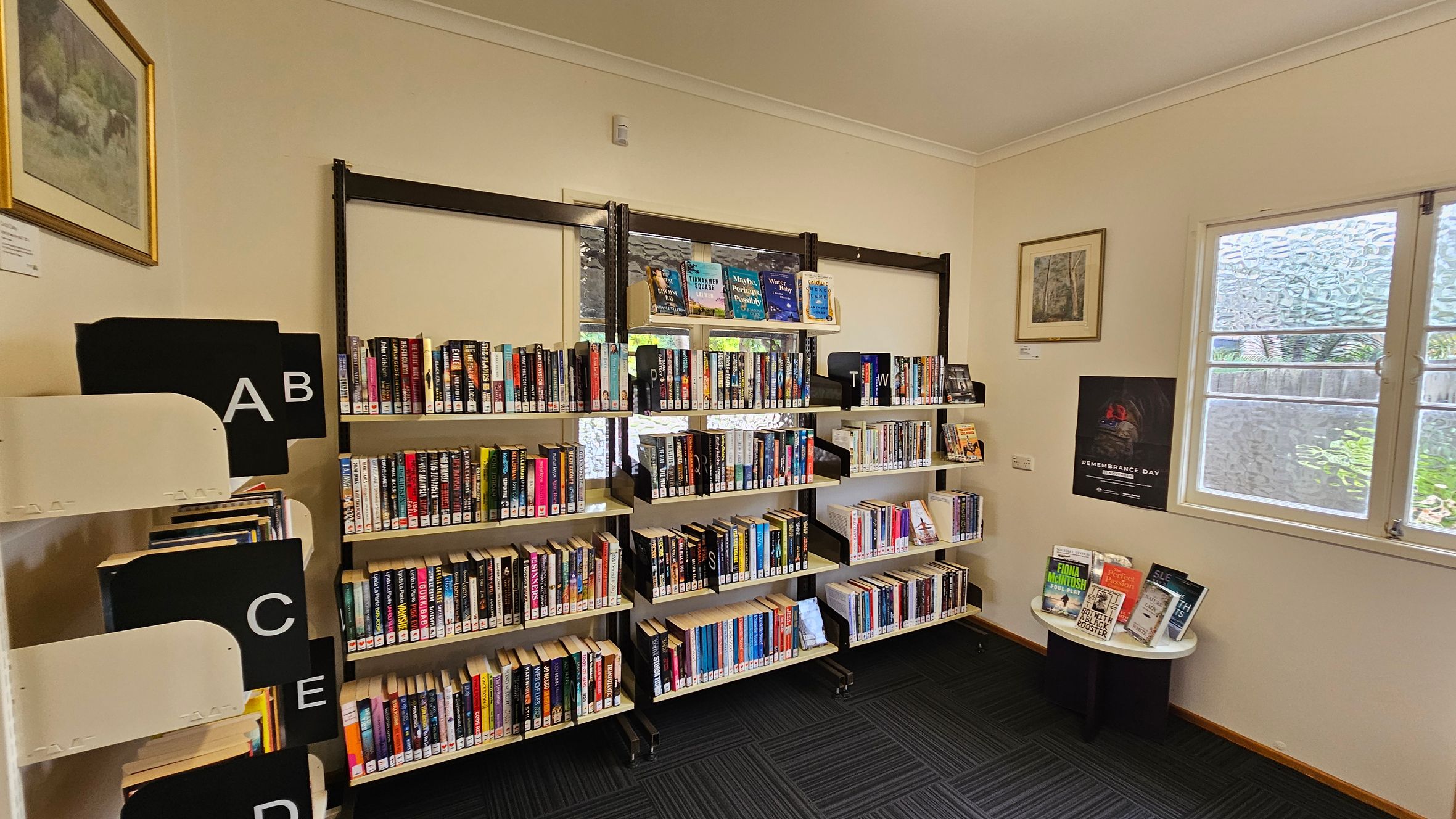 Bookshelves filled with books are against a cream-coloured wall. On the left, there are labelled sections with letters A to E. In the middle, three shelves hold books with dividers marked with letters. On the right, a small round table displays more books. Above it, a framed picture and a Remembrance Day poster are on the wall. A window with frosted glass is next to the table. The floor is carpeted with a dark pattern. Bookshelves filled with books are against a cream-coloured wall. On the left, there are labelled sections with letters A to E. In the middle, three shelves hold books with dividers marked with letters. On the right, a small round table displays more books. Above it, a framed picture and a Remembrance Day poster are on the wall. A window with frosted glass is next to the table. The floor is carpeted with a dark pattern.