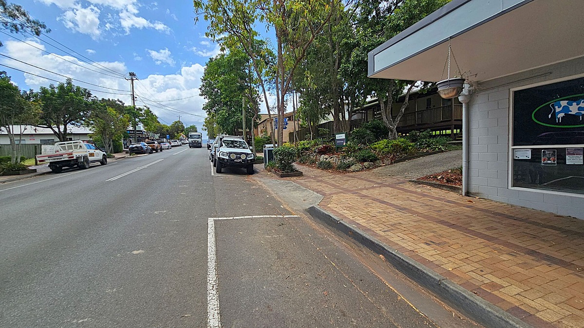 A street scene with a paved footpath on the right, bordered by a grey building with a hanging pot and a window displaying a cow graphic. Several cars are parked along the road, including a white vehicle in the foreground. Trees line the street, providing shade. On the left, more vehicles are parked, and a few buildings are visible in the distance. The road is marked with white lines, and the sky is partly cloudy. A street scene with a paved footpath on the right, bordered by a grey building with a hanging pot and a window displaying a cow graphic. Several cars are parked along the road, including a white vehicle in the foreground. Trees line the street, providing shade. On the left, more vehicles are parked, and a few buildings are visible in the distance. The road is marked with white lines, and the sky is partly cloudy.