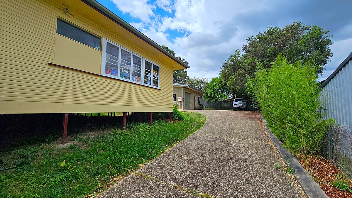 A yellow house on the left with large windows, elevated on stilts. A gravel driveway curves to the right, leading to a garage and a parked car. On the right, a tall green hedge runs alongside a metal fence. Trees are visible in the background. A yellow house on the left with large windows, elevated on stilts. A gravel driveway curves to the right, leading to a garage and a parked car. On the right, a tall green hedge runs alongside a metal fence. Trees are visible in the background.