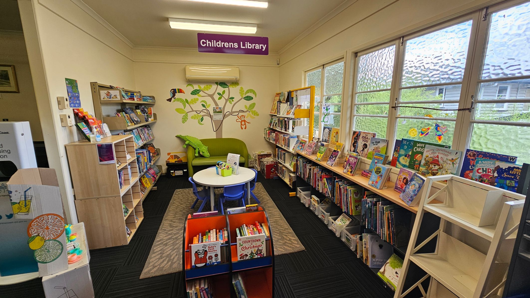 A library interior with a children's section on the left, featuring bookshelves filled with colourful books and a small play area with a lemonade stand. In the centre, a curved wooden counter displays brochures and a "Reservations" sign. Behind the counter, a computer and office supplies are visible. The room has fluorescent lighting and a ceiling fan. A wall clock is mounted on the right wall, and large windows provide natural light. The floor is carpeted with a dark pattern. A library interior with a children's section on the left, featuring bookshelves filled with colourful books and a small play area with a lemonade stand. In the centre, a curved wooden counter displays brochures and a "Reservations" sign. Behind the counter, a computer and office supplies are visible. The room has fluorescent lighting and a ceiling fan. A wall clock is mounted on the right wall, and large windows provide natural light. The floor is carpeted with a dark pattern.