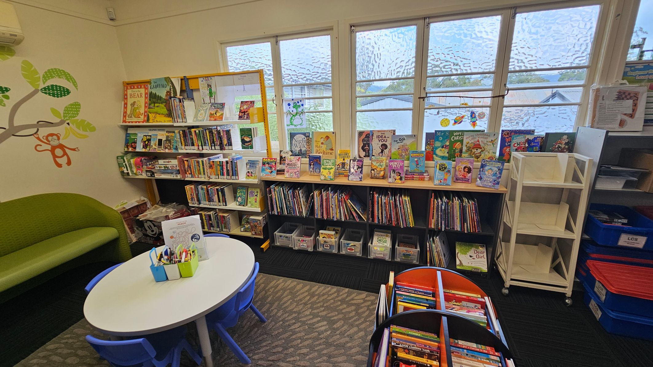 A cosy children's reading area with a green sofa on the left, decorated with a wall mural of a tree and a monkey. In the centre, a small round table with blue chairs is set up for activities, holding containers with pens. Bookshelves line the walls, filled with colourful children's books. Large windows in the background let in natural light. On the right, there are empty white shelves and blue storage bins. The floor is carpeted with a patterned rug. A cosy children's reading area with a green sofa on the left, decorated with a wall mural of a tree and a monkey. In the centre, a small round table with blue chairs is set up for activities, holding containers with pens. Bookshelves line the walls, filled with colourful children's books. Large windows in the background let in natural light. On the right, there are empty white shelves and blue storage bins. The floor is carpeted with a patterned rug.