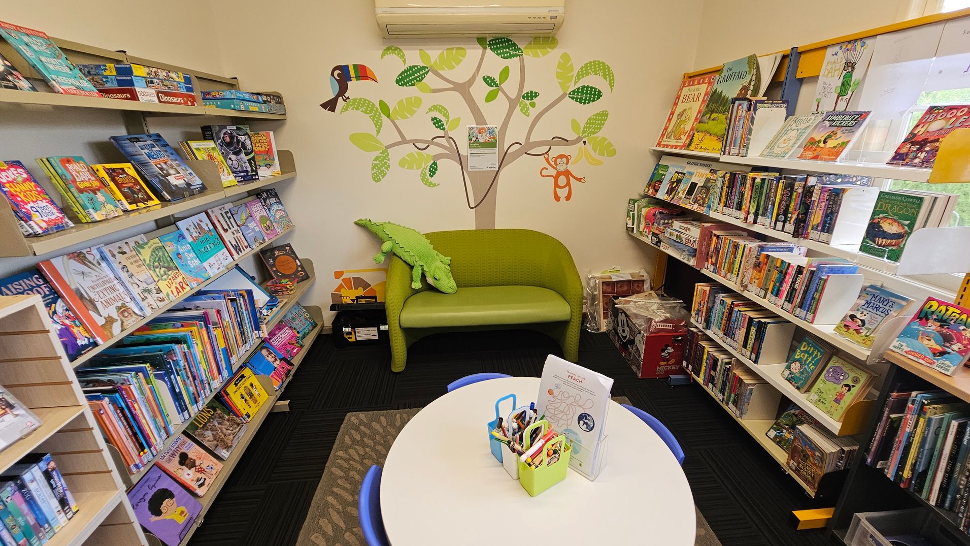 A cosy children's reading area with bookshelves on both sides filled with colourful books. In the centre, a small green sofa with a plush crocodile toy sits against a wall decorated with a tree mural featuring a toucan and a monkey. In the foreground, a round table with blue chairs holds containers of crayons and paper. The floor is carpeted in dark tones, and the ceiling is not visible. A cosy children's reading area with bookshelves on both sides filled with colourful books. In the centre, a small green sofa with a plush crocodile toy sits against a wall decorated with a tree mural featuring a toucan and a monkey. In the foreground, a round table with blue chairs holds containers of crayons and paper. The floor is carpeted in dark tones, and the ceiling is not visible.