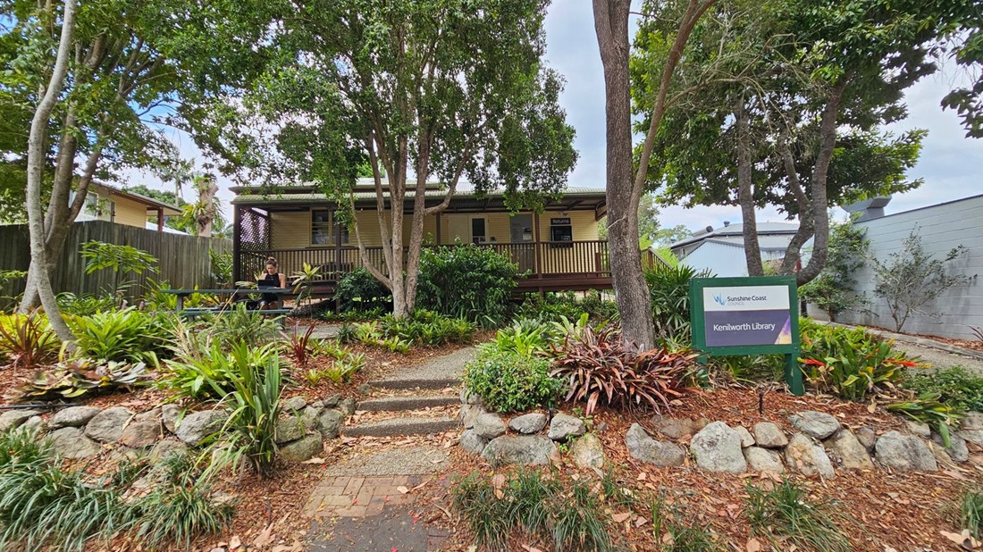 A small building with a wooden deck is surrounded by lush greenery and trees. A person sits at a picnic table on the left, using a laptop. A sign on the right reads "Kenilworth Library" with "Sunshine Coast" branding. The foreground features a stone-bordered garden with various plants and a pathway leading up to the building. A wooden fence is visible on the left, and a grey wall is on the right. A small building with a wooden deck is surrounded by lush greenery and trees. A person sits at a picnic table on the left, using a laptop. A sign on the right reads "Kenilworth Library" with "Sunshine Coast" branding. The foreground features a stone-bordered garden with various plants and a pathway leading up to the building. A wooden fence is visible on the left, and a grey wall is on the right.