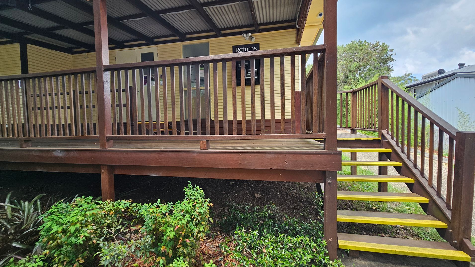 A wooden deck with a railing and a set of stairs leading up to it. The stairs have yellow edges for visibility. The building is painted yellow with a sign that reads "Returns" above a window. The roof is corrugated metal. In the foreground, there are green shrubs and plants. To the right, a path leads away from the stairs, and a grey building is partially visible in the background. A wooden deck with a railing and a set of stairs leading up to it. The stairs have yellow edges for visibility. The building is painted yellow with a sign that reads "Returns" above a window. The roof is corrugated metal. In the foreground, there are green shrubs and plants. To the right, a path leads away from the stairs, and a grey building is partially visible in the background.