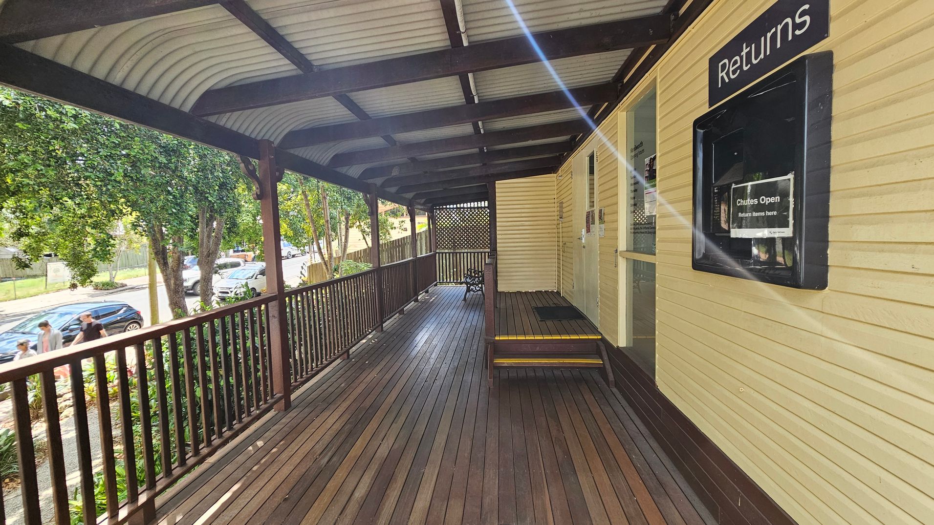 A wooden deck with a corrugated metal roof. On the right, a yellow wall features a "Returns" slot with a sign indicating "Chutes Open." The deck has a wooden railing, and a small set of steps leads to a door. On the left, trees and parked cars line the street, with a few people walking on the pavement. The floor is made of wooden planks. A wooden deck with a corrugated metal roof. On the right, a yellow wall features a "Returns" slot with a sign indicating "Chutes Open." The deck has a wooden railing, and a small set of steps leads to a door. On the left, trees and parked cars line the street, with a few people walking on the pavement. The floor is made of wooden planks.
