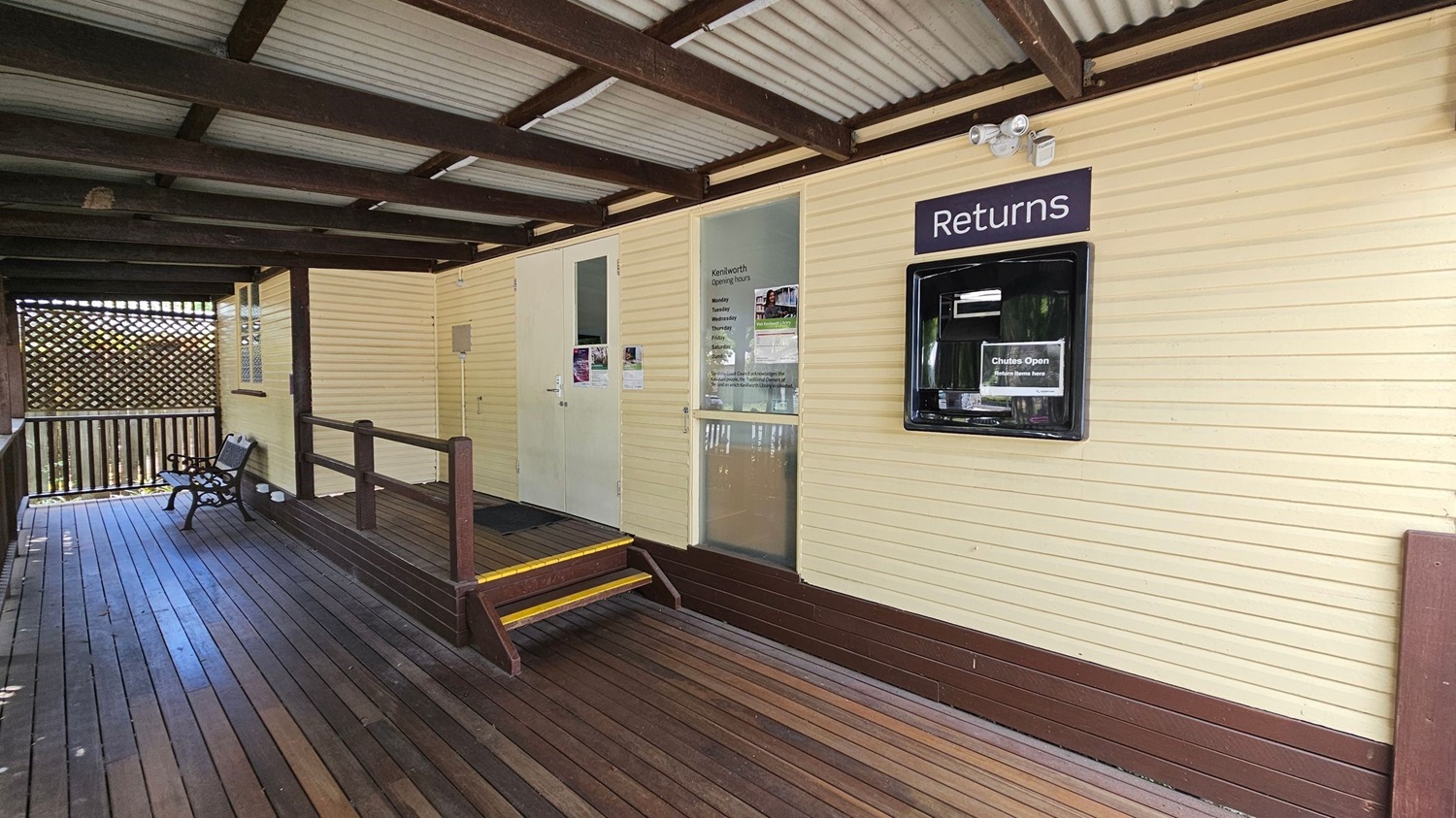 A wooden deck with a bench on the left side, leading to a building with cream-coloured walls. The building has a "Returns" slot on the right, and a glass door with opening hours listed. A small set of steps with yellow edges leads up to the door. The ceiling is made of corrugated metal with wooden beams. A lattice screen is visible in the background on the left. A wooden deck with a bench on the left side, leading to a building with cream-coloured walls. The building has a "Returns" slot on the right, and a glass door with opening hours listed. A small set of steps with yellow edges leads up to the door. The ceiling is made of corrugated metal with wooden beams. A lattice screen is visible in the background on the left.