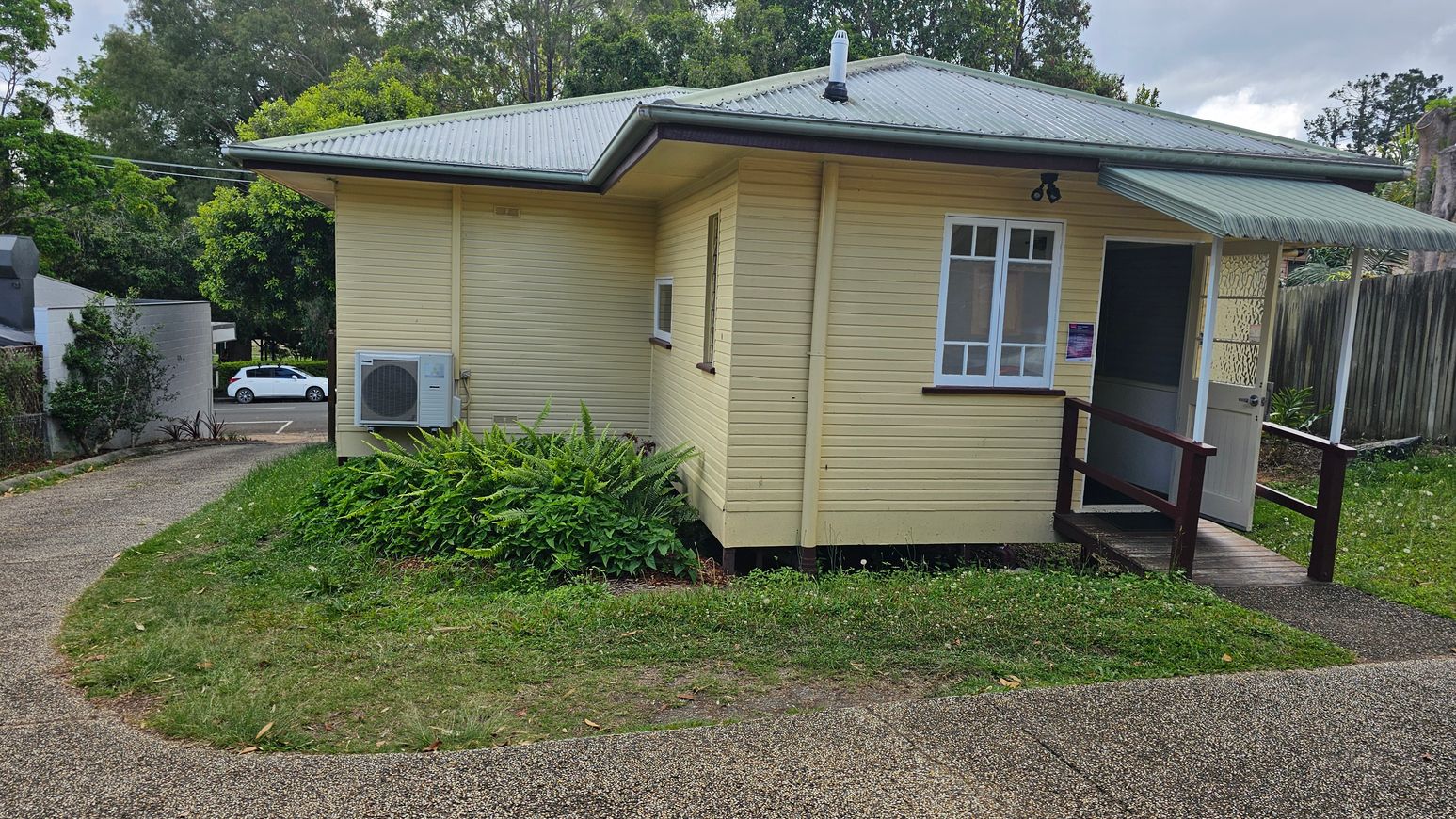 A small, single-storey yellow house with a grey metal roof. On the right, there's a partially open door with a small awning above it. A window is next to the door. An air conditioning unit is mounted on the left side of the house. The house is surrounded by grass and ferns. A paved path curves around the house, leading to a road where a white car is parked. Trees and a wooden fence are in the background. A small, single-storey yellow house with a grey metal roof. On the right, there's a partially open door with a small awning above it. A window is next to the door. An air conditioning unit is mounted on the left side of the house. The house is surrounded by grass and ferns. A paved path curves around the house, leading to a road where a white car is parked. Trees and a wooden fence are in the background.