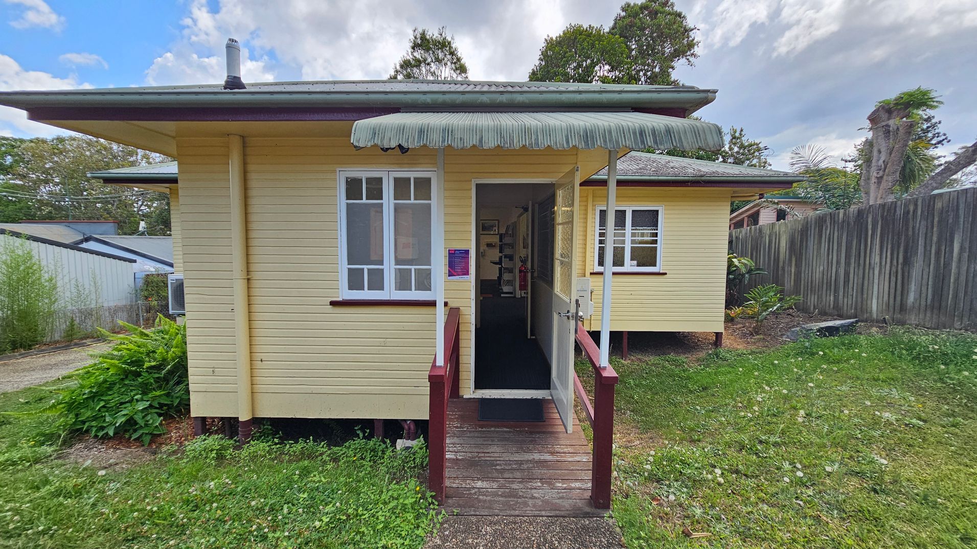 A small, single-storey yellow wooden house with a green corrugated awning above the entrance. The entrance has a wooden ramp with red railings leading to an open door. Two white-framed windows are visible on either side of the door. The house is elevated slightly on stilts. To the right, there's a wooden fence and some greenery. The left side shows a neighbouring building and more plants. The grass in the foreground is patchy with some weeds. A small, single-storey yellow wooden house with a green corrugated awning above the entrance. The entrance has a wooden ramp with red railings leading to an open door. Two white-framed windows are visible on either side of the door. The house is elevated slightly on stilts. To the right, there's a wooden fence and some greenery. The left side shows a neighbouring building and more plants. The grass in the foreground is patchy with some weeds.