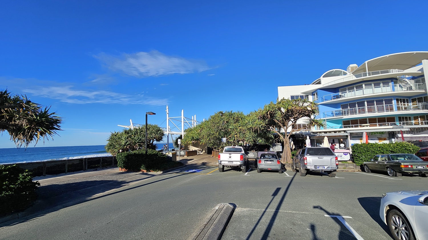 On the left, there is a coastal walkway lined with bushes and a tree, leading to the ocean. In the middle, there is a pathway with people walking, flanked by parked cars and trees. On the right, there is a multi-storey building with balconies and glass railings, adjacent to more parked cars. The foreground shows a carpark with multiple vehicles parked. The floor is a mix of asphalt and concrete. On the left, there is a coastal walkway lined with bushes and a tree, leading to the ocean. In the middle, there is a pathway with people walking, flanked by parked cars and trees. On the right, there is a multi-storey building with balconies and glass railings, adjacent to more parked cars. The foreground shows a carpark with multiple vehicles parked. The floor is a mix of asphalt and concrete.