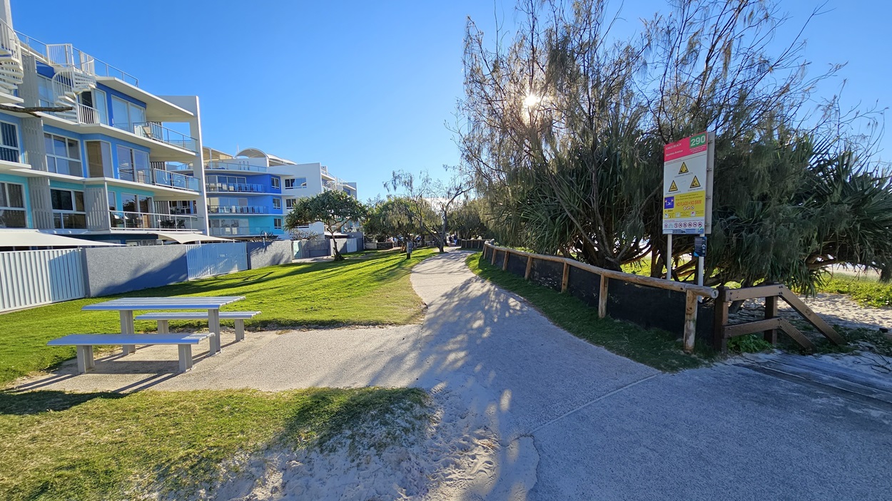 On the left, there is a grey picnic table on a grassy area, with a multi-storey building featuring balconies and glass railings behind it. In the middle, there is a winding concrete pathway bordered by grass, leading towards trees and more buildings in the background. On the right, there is a wooden fence with a large sign displaying warnings and information, next to dense bushes and trees. The floor is a mix of concrete and sand and there is a picnic table visible on the left. On the left, there is a grey picnic table on a grassy area, with a multi-storey building featuring balconies and glass railings behind it. In the middle, there is a winding concrete pathway bordered by grass, leading towards trees and more buildings in the background. On the right, there is a wooden fence with a large sign displaying warnings and information, next to dense bushes and trees. The floor is a mix of concrete and sand and there is a picnic table visible on the left.