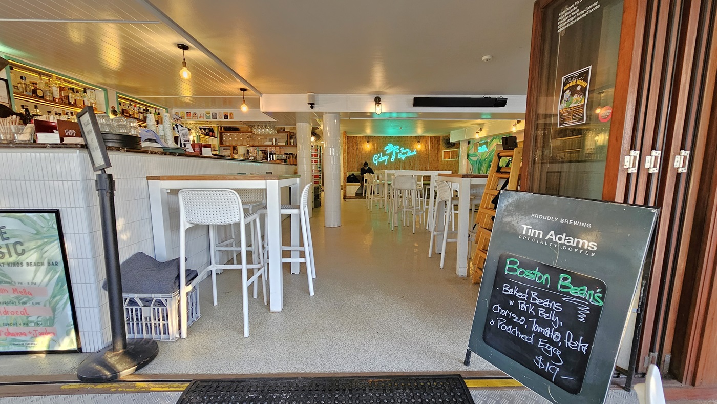 On the left, there is a white display freezer with an owl figurine on top and a glass display case containing food items. In the middle, there is an open entrance leading into a café with white tables and chairs, a counter with stools, and a neon sign on the back wall. On the right, there are more white tables and chairs, a mural of a parrot on the wall, and a wooden partition. The floor is concrete, and the ceiling has exposed wooden beams and white panels. On the left, there is a white display freezer with an owl figurine on top and a glass display case containing food items. In the middle, there is an open entrance leading into a café with white tables and chairs, a counter with stools, and a neon sign on the back wall. On the right, there are more white tables and chairs, a mural of a parrot on the wall, and a wooden partition. The floor is concrete, and the ceiling has exposed wooden beams and white panels.
