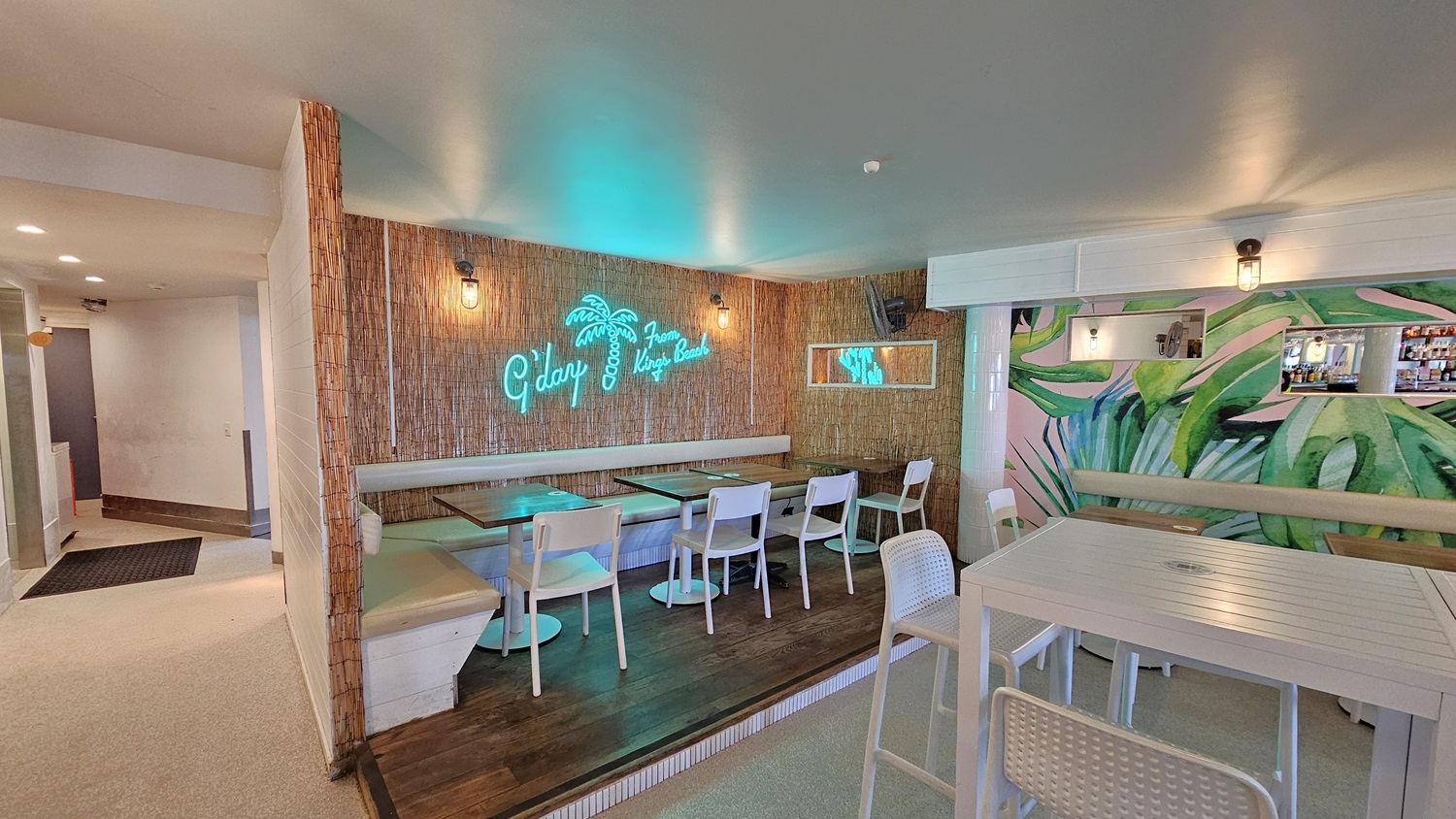 On the left, there is a bamboo wall with a neon sign reading "G'day From Kings Beach" above a seating area with brown tables and beige cushioned benches. There is a wooden platform with additional seating and a small mirror on the wall. On the right, there is a mural of green leaves on the wall, a bar counter with stools, and shelves with bottles. The floor is light-coloured and the ceiling is white with recessed lighting.A corridor leading to an area with white walls is visible on the left. On the left, there is a bamboo wall with a neon sign reading "G'day From Kings Beach" above a seating area with brown tables and beige cushioned benches. There is a wooden platform with additional seating and a small mirror on the wall. On the right, there is a mural of green leaves on the wall, a bar counter with stools, and shelves with bottles. The floor is light-coloured and the ceiling is white with recessed lighting.A corridor leading to an area with white walls is visible on the left.