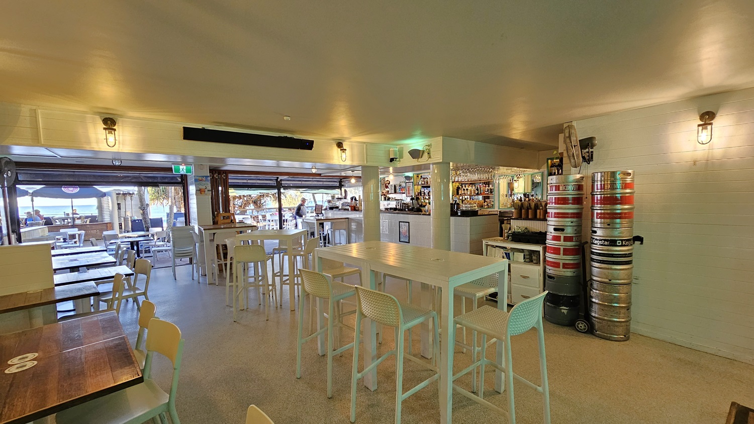 On the left, there are white tables and chairs near a wooden wall with a window. In the middle, there are high white tables with bar stools, and a bar counter with various bottles and glasses. In the background, there is an open area with more tables and chairs, and a view of the beach. On the right, there are stacked beer kegs and a white brick wall. The floor is light-coloured concrete, and the ceiling is white with recessed lighting and wall-mounted lamps. On the left, there are white tables and chairs near a wooden wall with a window. In the middle, there are high white tables with bar stools, and a bar counter with various bottles and glasses. In the background, there is an open area with more tables and chairs, and a view of the beach. On the right, there are stacked beer kegs and a white brick wall. The floor is light-coloured concrete, and the ceiling is white with recessed lighting and wall-mounted lamps.