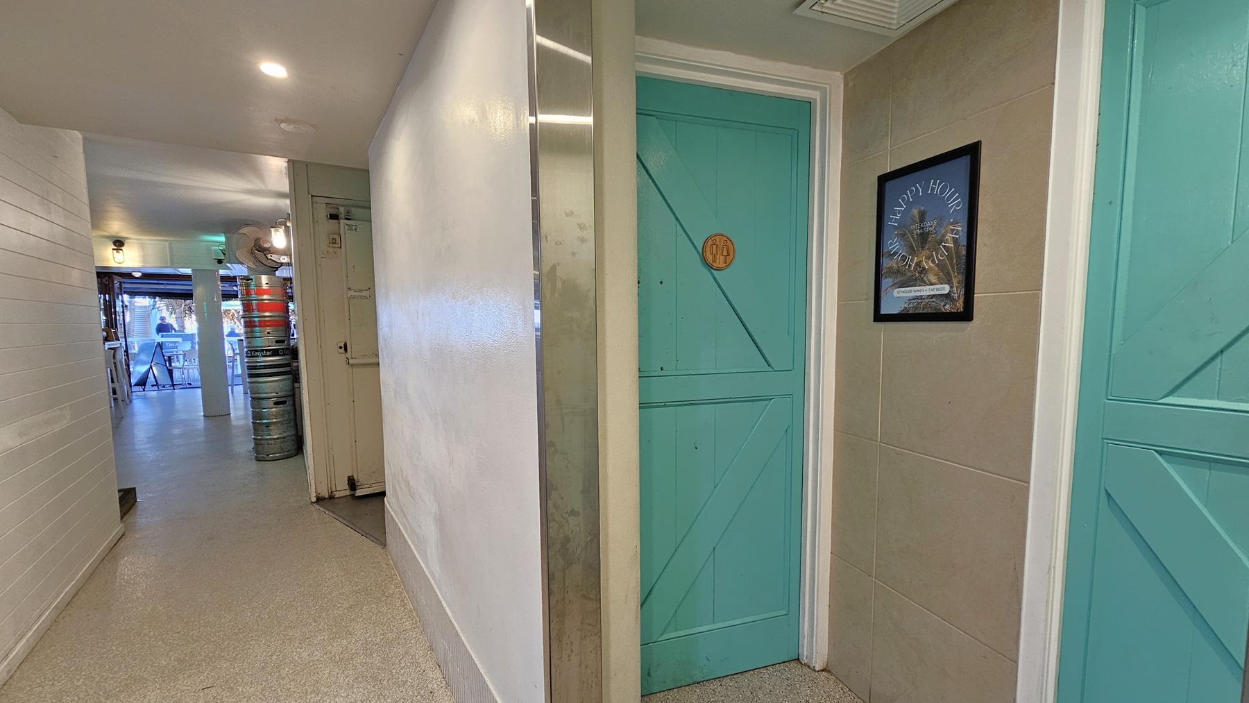 On the left, there is a hallway with white walls and a light-coloured floor, leading to an open area with stacked beer kegs and a view of tables and chairs. In the middle, there is a white wall with a narrow vertical mirror. On the right, there are two turquoise doors with wooden panels, one with a "WC" sign and the other with a framed picture of a beach scene. The floor is light-coloured concrete, and the ceiling is white with recessed lighting. On the left, there is a hallway with white walls and a light-coloured floor, leading to an open area with stacked beer kegs and a view of tables and chairs. In the middle, there is a white wall with a narrow vertical mirror. On the right, there are two turquoise doors with wooden panels, one with a "WC" sign and the other with a framed picture of a beach scene. The floor is light-coloured concrete, and the ceiling is white with recessed lighting.