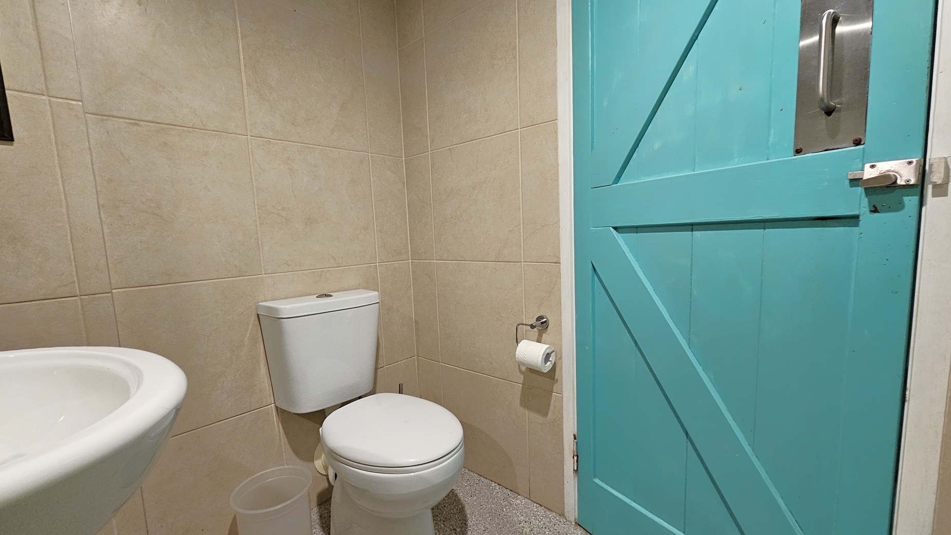 On the left, there is a white sink with a round basin and a beige tiled wall. In the middle, there is a white toilet with a toilet roll holder mounted on the wall beside it. On the right, there is a closed turquoise door with a metal handle and latch. The floor is speckled grey tiles. On the left, there is a white sink with a round basin and a beige tiled wall. In the middle, there is a white toilet with a toilet roll holder mounted on the wall beside it. On the right, there is a closed turquoise door with a metal handle and latch. The floor is speckled grey tiles.