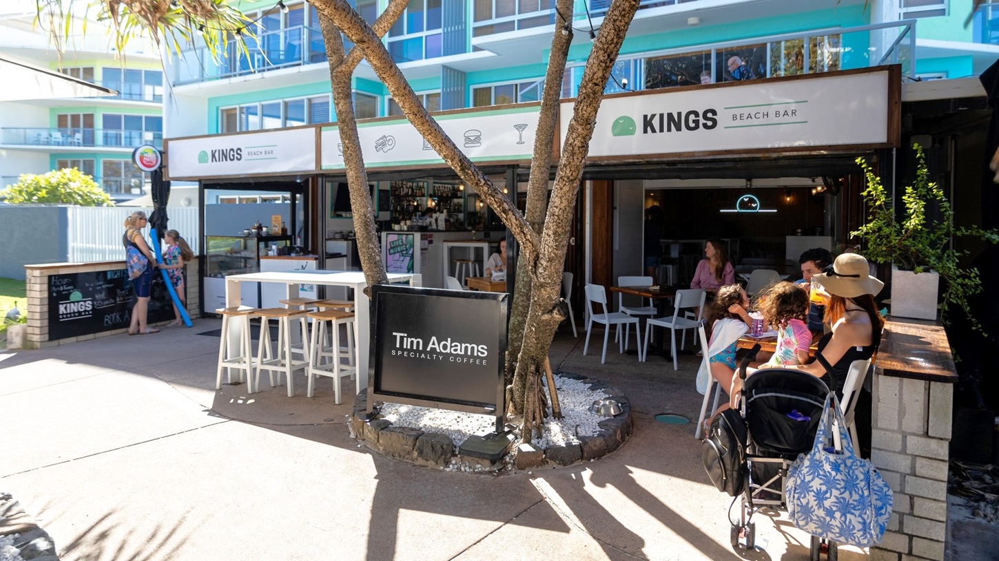 On the left, there is a white fence and a sign for Kings Beach Bar, with people walking past. In the middle, there is an outdoor seating area with white tables and chairs, some occupied by people, and a bar counter with stools. A tall palm tree with a slender trunk bearing a sign "Tim Adams" stands in the middle, bordered by stones and pebbles. On the right, a family with a pram sits at a table near another smaller tree. The background features a multi-storey building with blue balconies. On the left, there is a white fence and a sign for Kings Beach Bar, with people walking past. In the middle, there is an outdoor seating area with white tables and chairs, some occupied by people, and a bar counter with stools. A tall palm tree with a slender trunk bearing a sign "Tim Adams" stands in the middle, bordered by stones and pebbles. On the right, a family with a pram sits at a table near another smaller tree. The background features a multi-storey building with blue balconies.