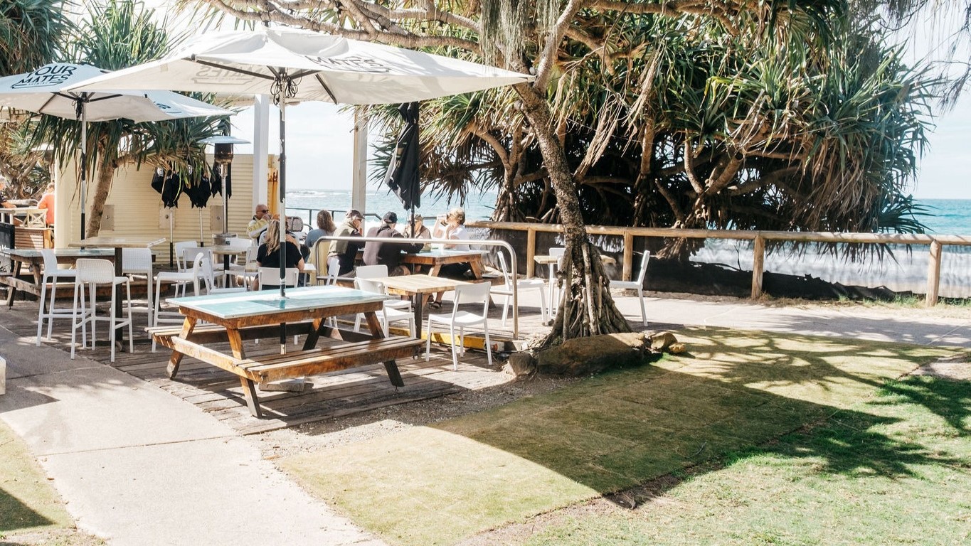 On the left, there are white umbrellas over tables and chairs, with people sitting and a bar counter with stools nearby. In the middle, there is a large tree with a thick trunk and sprawling branches, surrounded by more tables and chairs, including picnic tables. On the right, there is a wooden fence with dense foliage and the ocean visible beyond it. The floor is a mix of concrete and grass, and the ocean is visible on the right. On the left, there are white umbrellas over tables and chairs, with people sitting and a bar counter with stools nearby. In the middle, there is a large tree with a thick trunk and sprawling branches, surrounded by more tables and chairs, including picnic tables. On the right, there is a wooden fence with dense foliage and the ocean visible beyond it. The floor is a mix of concrete and grass, and the ocean is visible on the right.
