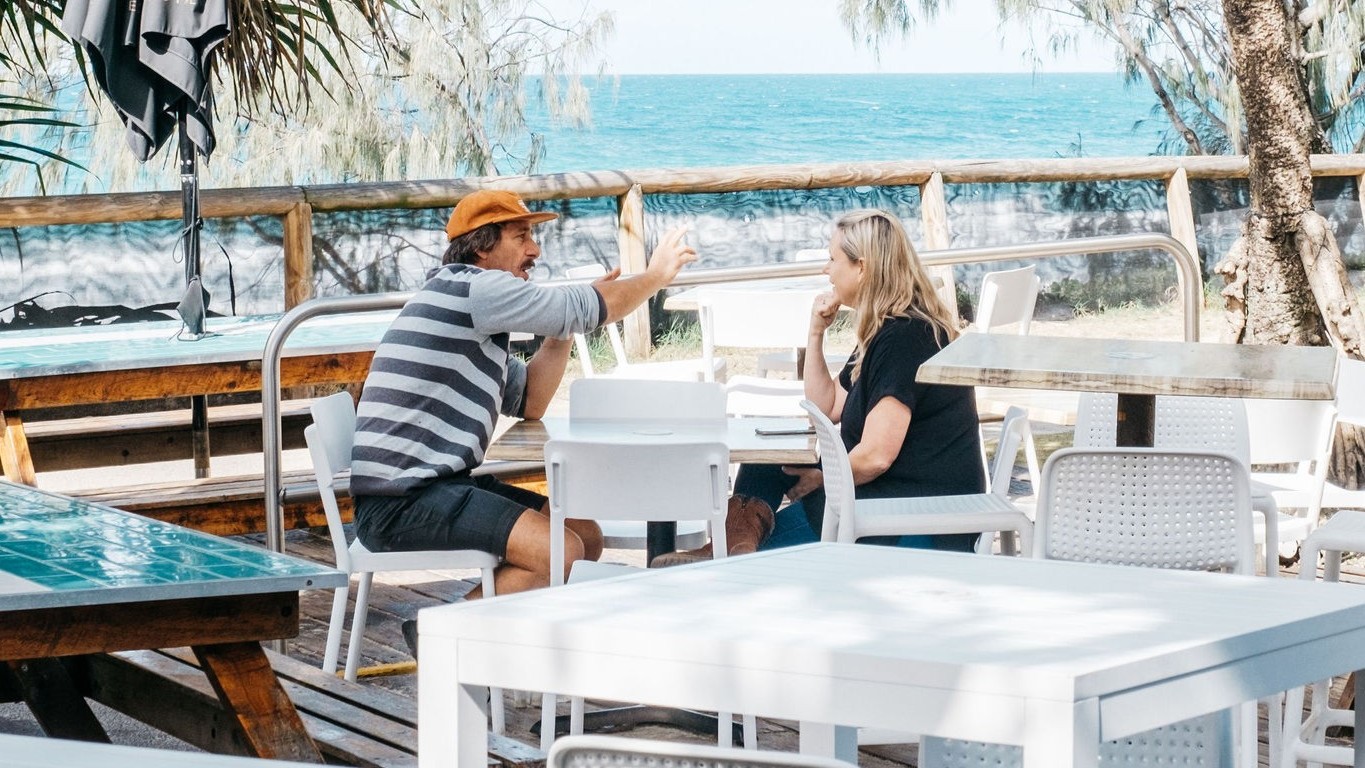 There are white tables and chairs with a man in a striped shirt and orange hat sitting and gesturing. In the middle, a woman with blonde hair in a black shirt sits across from him at a white table. On the right, there is a tree with a thick trunk and more white tables and chairs. The background features a wooden fence and the ocean beyond it. The floor is wooden decking, and a folded umbrella shade is positioned on the left. There are white tables and chairs with a man in a striped shirt and orange hat sitting and gesturing. In the middle, a woman with blonde hair in a black shirt sits across from him at a white table. On the right, there is a tree with a thick trunk and more white tables and chairs. The background features a wooden fence and the ocean beyond it. The floor is wooden decking, and a folded umbrella shade is positioned on the left.