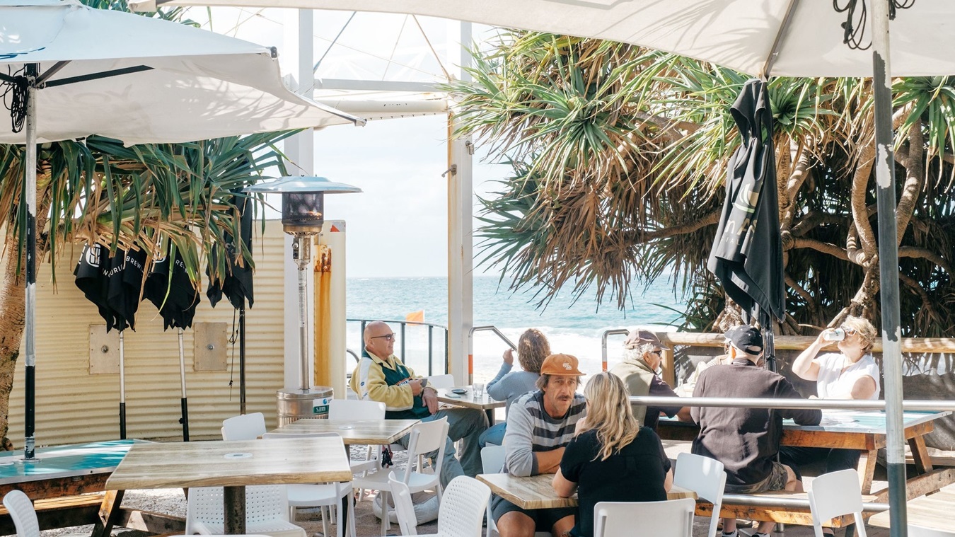 On the left, there are white tables and chairs under a large umbrella, with a man in a yellow jacket sitting at one of the tables. In the middle, a man in an orange hat and a woman with blonde hair sit at a white table, facing each other. On the right, there is a large tree with dense foliage, more tables and chairs, and people sitting and talking. The background features a wooden fence and the ocean beyond it. The floor is wooden decking. On the left, there are white tables and chairs under a large umbrella, with a man in a yellow jacket sitting at one of the tables. In the middle, a man in an orange hat and a woman with blonde hair sit at a white table, facing each other. On the right, there is a large tree with dense foliage, more tables and chairs, and people sitting and talking. The background features a wooden fence and the ocean beyond it. The floor is wooden decking.