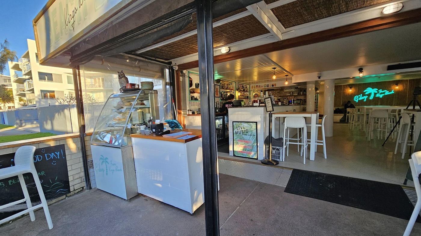 On the left, there is an outdoor area with a white display freezer and a glass display case containing food items. In the middle, there is an open entrance leading into a café with white tables and chairs, a counter with stools, and a neon sign on the back wall. On the right, inside the café, there are more white tables and chairs, and a bar counter with various items. The floor is concrete, and the ceiling has exposed wooden beams and white panels. On the left, there is an outdoor area with a white display freezer and a glass display case containing food items. In the middle, there is an open entrance leading into a café with white tables and chairs, a counter with stools, and a neon sign on the back wall. On the right, inside the café, there are more white tables and chairs, and a bar counter with various items. The floor is concrete, and the ceiling has exposed wooden beams and white panels.